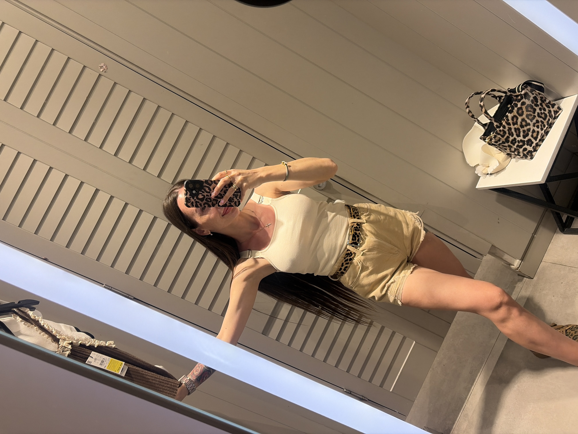 A woman is taking a mirror selfie in a fitting room. She is wearing a white tank top, khaki shorts, and a leopard print belt. She is holding a phone with a leopard print case and has several rings on her fingers. In the background, there are slatted doors and a shelf with a white hat and a leopard print bag. The environment appears to be a retail store fitting room in Saint Petersburg, Russia.