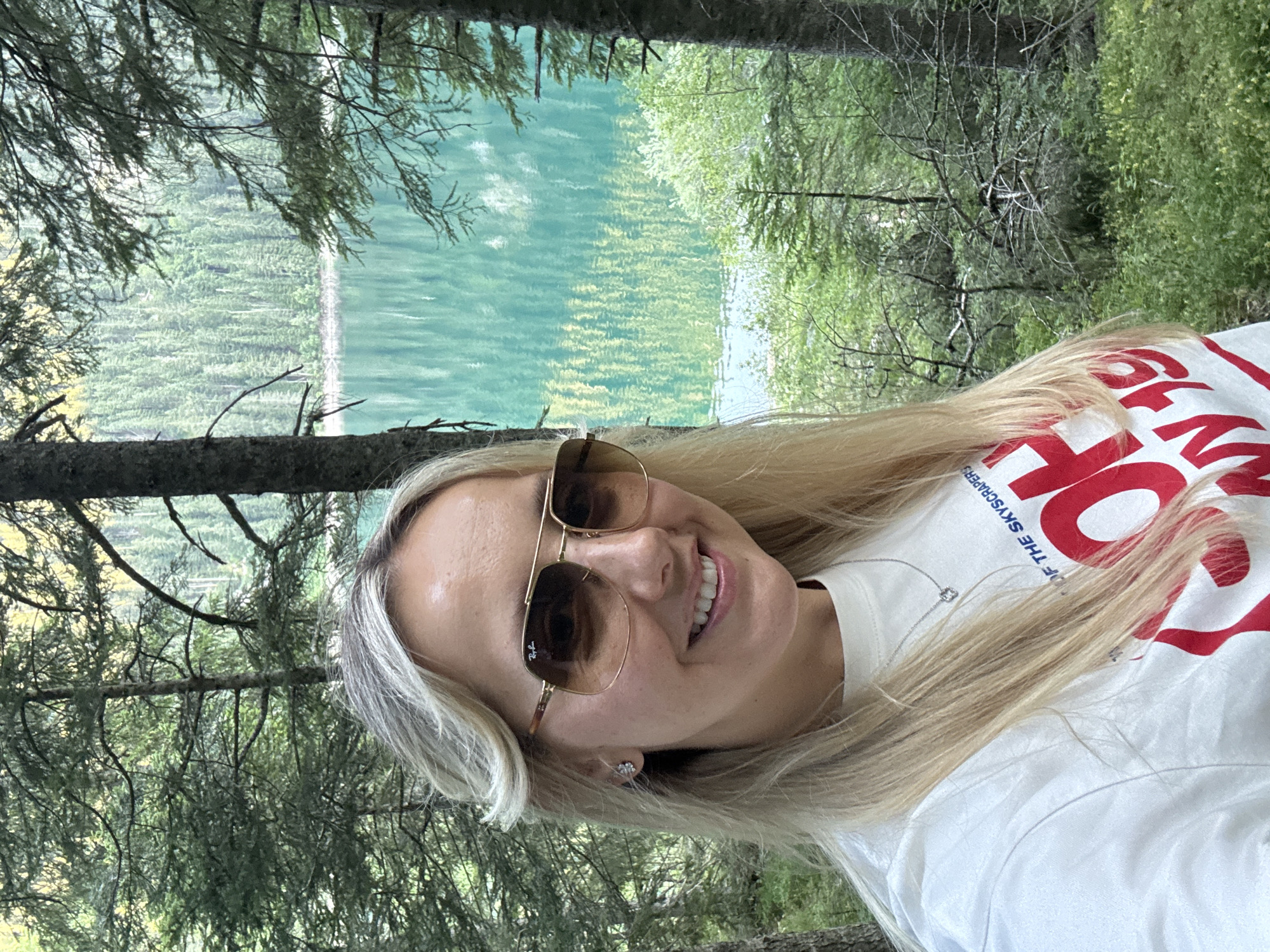 A woman with blonde hair and sunglasses is taking a selfie outdoors. She is wearing a white t-shirt with red text. She is smiling and looking at the camera. The background shows a lake surrounded by trees. The scene appears to be in a natural setting, possibly a park or forest. The location is identified as Pitomine, Montenegro.