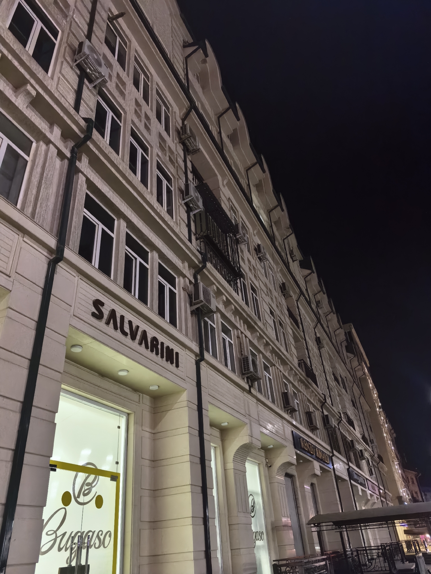 An exterior, low-angle, night-time shot showcases the facade of a modern, cream-colored building in Fergana City, Uzbekistan. The building features multiple stories with numerous rectangular windows, many equipped with air conditioning units. Distinct signage identifies businesses on the ground floor, including "SALVARINI" in large, dark letters above a storefront with glass doors displaying the "Bugaso" logo. The architecture exhibits classical detailing with horizontal banding and decorative elements along the roofline. The street is dimly lit, with the building's internal lighting casting a warm glow onto the pavement and illuminating the storefronts. The sky is a deep, inky black, suggesting the late hour.