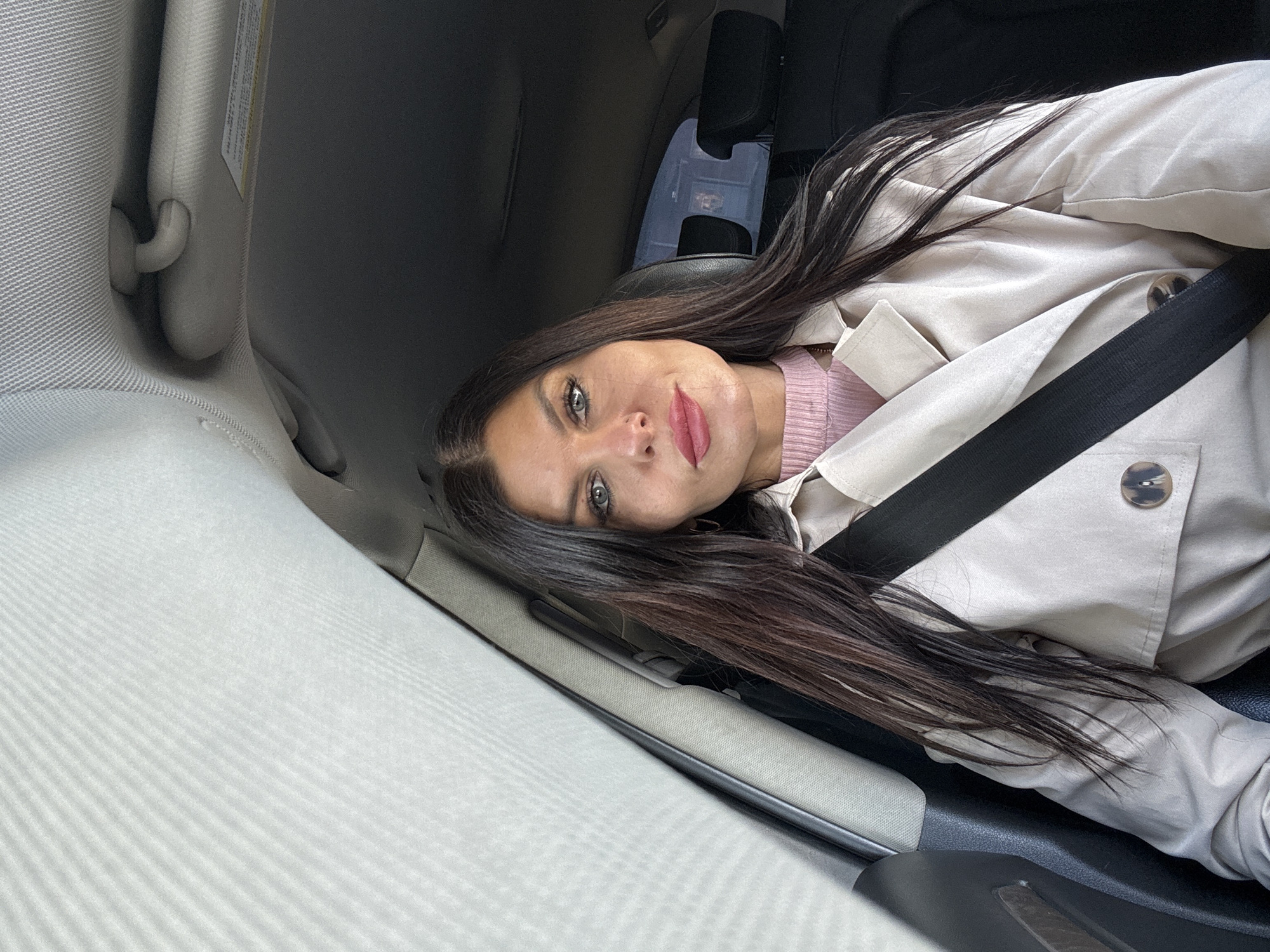 A woman with long brown hair, wearing a pink top and a beige trench coat, is secured in a car seat with a black seatbelt. The interior of the car is visible, with a light gray sun visor and upholstery. The location in the city of Chaikovsky, Russia is not discernible from the image.