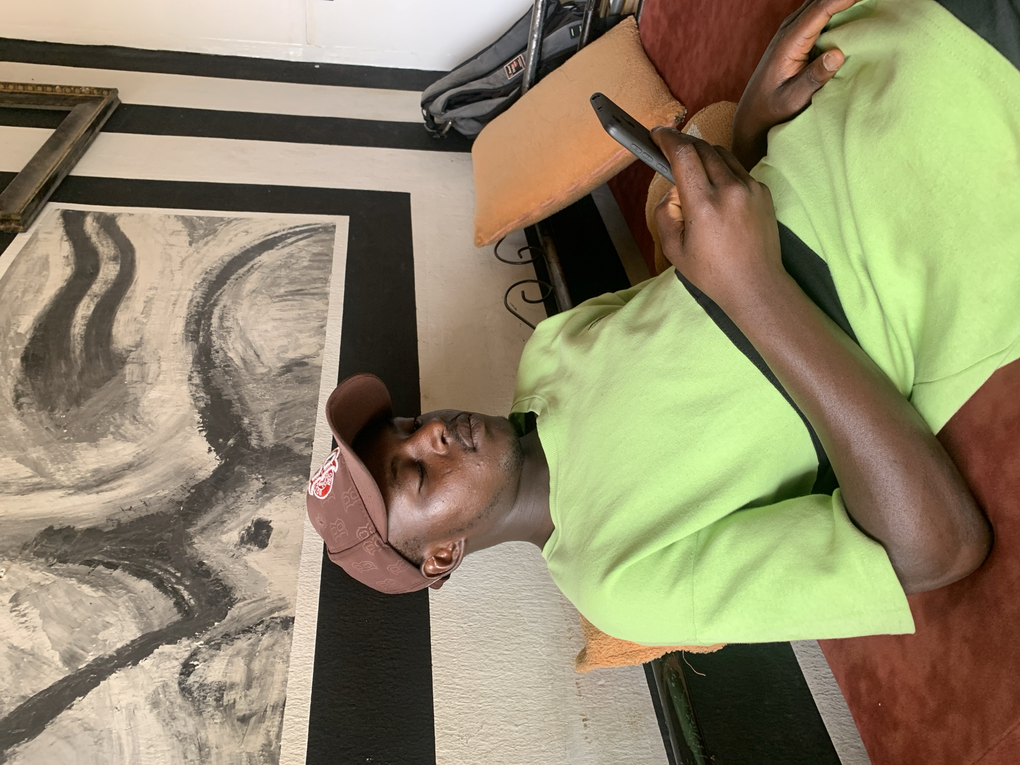 A dark-skinned man is depicted reclining indoors, engrossed in his smartphone. He wears a light green short-sleeved shirt and a brown baseball cap featuring a red and white logo, which appears to include the letter 'B' or 'R' and other design elements. His eyes are closed or cast downwards, suggesting a moment of focus on the device or relaxation.
He is situated on a reddish-brown cushioned piece of furniture, likely a sofa or chair, with an orange cushion supporting him. The background features a striking black and white striped pattern on what appears to be a wall or floor. Adjacent to him, an abstract painting in black, white, and grey tones rests on this striped surface, alongside an ornate, empty gold picture frame. A black bag or backpack with a faint, indiscernible white logo is visible near the orange cushion. The scene conveys a casual and relaxed atmosphere within a brightly lit interior space in Jos, Nigeria, during the daytime.