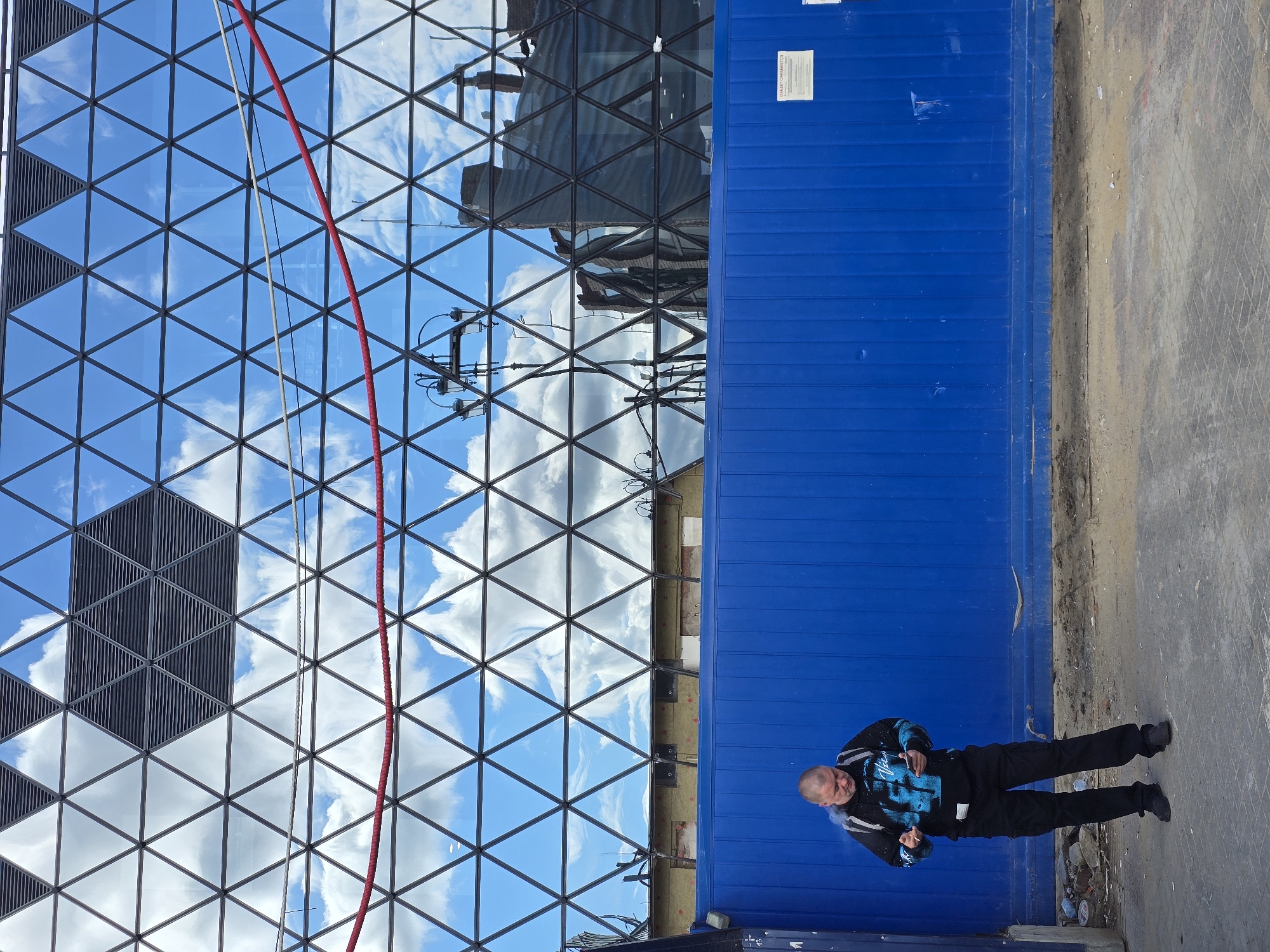 A middle-aged man wearing a black and blue sweatshirt and black cargo pants stands in front of a bright blue metal fence. He has a backpack on and is looking down at his phone. The background is a large, modern building with a triangular glass facade reflecting a blue sky with white clouds. A red cord runs horizontally across the glass facade. The ground in front of the fence is dirt and gravel, with some debris scattered around. The overall scene appears to be an urban construction site. The building's architecture suggests it may be the "House of the Moscow Electronic Music" (also known as Domkult) located on Bolotnaya Embankment in Moscow, Russia.
