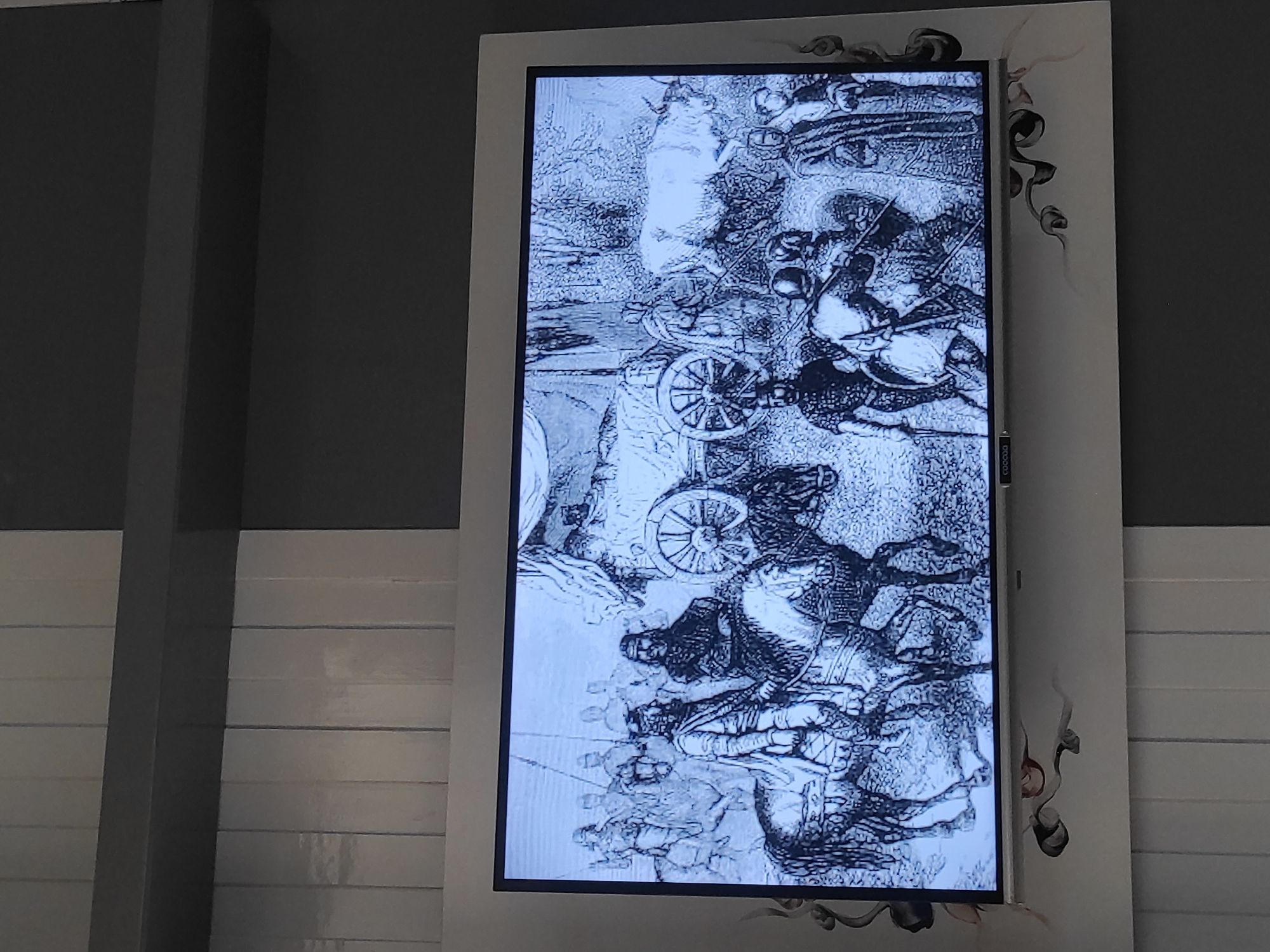 The image displays a television screen showing a black and white historical engraving. The engraving depicts a scene with several people on horseback and on foot, along with a horse-drawn wagon and a cow. The people are dressed in what appears to be historical attire, with some wearing armor and helmets. The setting seems to be outdoors, possibly in a rural or historical landscape with trees and foliage suggested in the background. The engraving is detailed, showing the textures of clothing, animals, and the rough terrain. This image is displayed on a modern flat-screen television mounted on a wall that has vertical white panels and a dark gray horizontal band above it. Decorative dark swirls are present on the wall surrounding the television. This scene is located in the city of Sumedang, Indonesia, specifically in a public area or establishment where this display is situated.