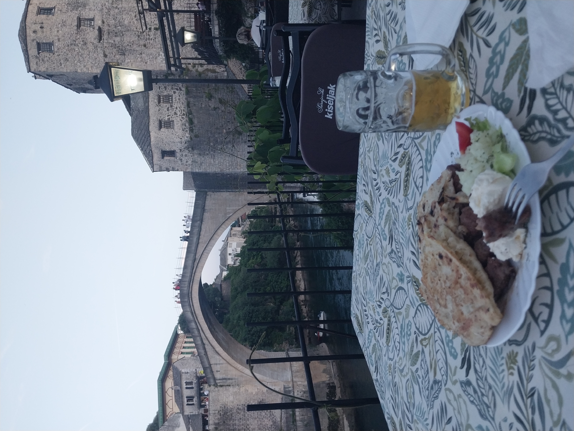 This media file captures an outdoor dining scene in Mostar, Bosnia and Herzegovina, featuring the iconic Stari Most (Old Bridge) in the background. The image is taken from a cafe or restaurant terrace, with a table set in the foreground.
In the immediate foreground, a patterned tablecloth (featuring green and blue leaf motifs) covers a table. On the table, there's a white plate holding a meal, likely local cuisine, consisting of meat (possibly ćevapi), flatbread (lepina or somun), a dollop of a white sauce or cream (possibly kaymak or pavlaka), and a small side salad with lettuce and a slice of tomato. A metal fork is visible, resting on the plate. Next to the plate, a large, clear glass beer mug, partially filled with a golden beverage (presumably beer), sits on the tablecloth.
The midground features several dark-colored outdoor chairs, some of which bear the white text "Sarajevsko Kiseljak," indicating a brand of mineral water, possibly sponsoring the seating. Behind the chairs, a metal railing and some green foliage can be seen. A street lamp, glowing brightly, stands prominently to the left of the chairs, suggesting it's either dusk or early evening, or perhaps still daytime but the light is on.
The background showcases the impressive Stari Most arching over the Neretva River. Numerous people are visible walking across the bridge. Historic stone buildings line the riverbanks and surround the bridge, characteristic of Mostar's old town architecture. The sky above is clear and bright, consistent with late afternoon or early evening light, given the artificial light from the street lamp. The overall setting evokes a relaxed atmosphere, likely enjoyed by tourists or locals dining with a scenic view.