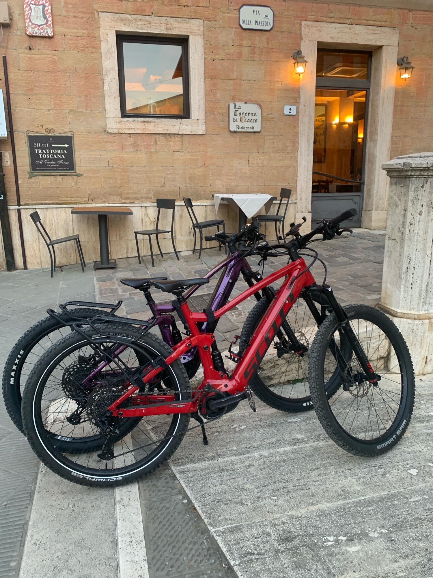 Two electric bicycles, a red one in the foreground and a purple one behind it, are parked on a paved walkway outside a building with a light brown brick facade. The red bicycle is a Scott brand, identifiable by the logo on its frame. Both bikes have front and rear suspension and disc brakes. The red bike has a rear rack with a black bag attached. A dark-colored chair is visible to the left of the bicycles. Further left, a sign with a shield and the word "Castello" is mounted on the wall, and below it, a smaller black sign with white text reads "50 mt Trattoria Toscana Al Vecchio Forno".
Above and to the right of the entrance to the building, two street signs are visible: one reading "VIA DELLA PIAZZOLA" and another with the number "2". A sign for "La Taverna di Ciacco Ristorante" is also present. The building's entrance is a doorway with a glass door and two ornate outdoor lights flanking it. Inside the building, visible through the glass, are illuminated tables and chairs. Two small tables with white tablecloths and chairs are set up outside the establishment to the left of the entrance. The architectural style of the building, including the stonework around the window and door, suggests an Italian setting.