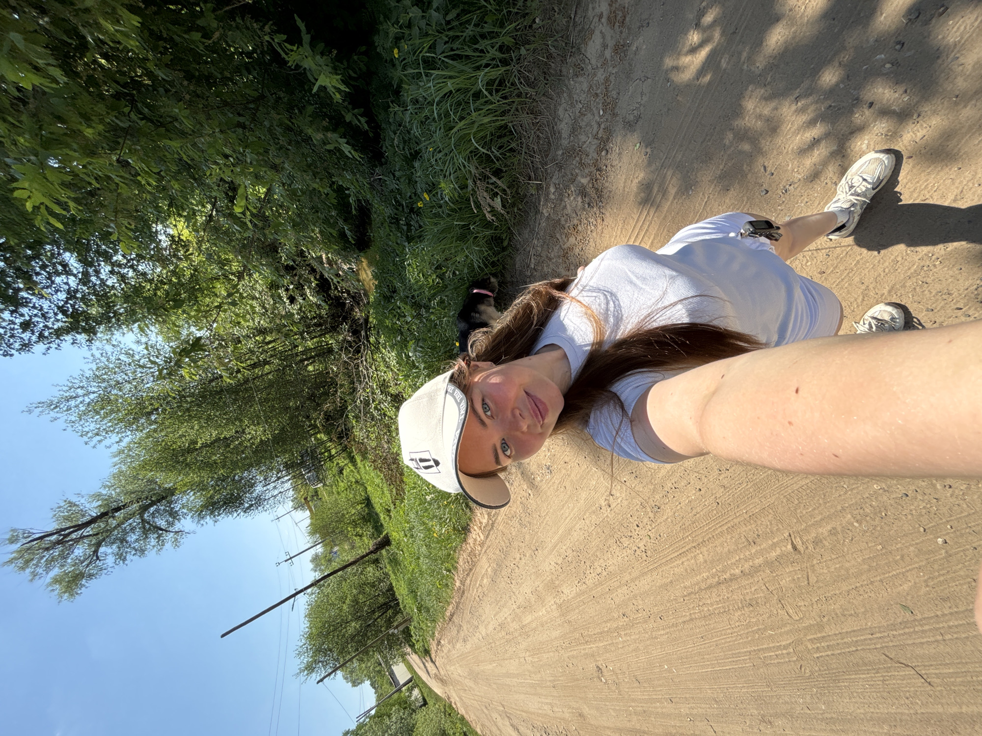A woman with long brown hair, wearing a white t-shirt, white shorts, a white cap with a black logo, and white sneakers, is seen on a dirt road in Malyi Bukor, Russia. She is holding the camera at arm's length, capturing a selfie. A dog is visible in the background, partially obscured by vegetation. The road is unpaved, and lush green vegetation borders the road on both sides. Overhead, trees and a clear blue sky are visible. Utility poles are on the left side of the road.
