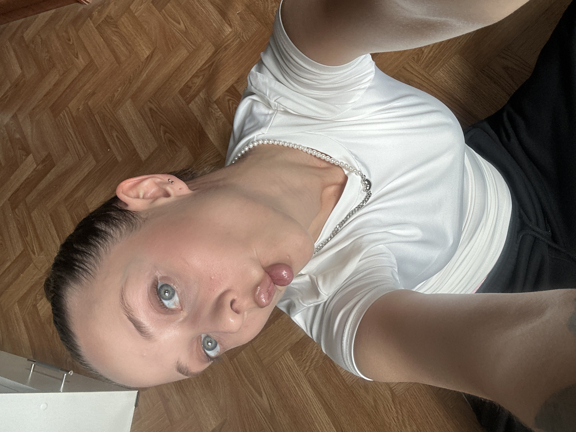 The image shows a close-up, overhead shot of a person lying down on a wooden floor. The person is wearing a white shirt with ruched detailing and a pearl necklace. They have blue eyes and their tongue is sticking out, with a slight bulge. Their hair is dark and pulled back. To their right, a portion of black fabric, likely pants, is visible.
The scene appears to be indoors, as suggested by the wooden floor and what looks like the edge of a white piece of furniture or shelving unit in the lower left corner. The lighting is natural and appears to be coming from above, creating some shadows on the person's face.
The activity taking place is a selfie, with the camera positioned above and looking down at the person. The emotion conveyed is playful or mischievous, due to the tongue-out pose. There is no visible text in the image. The location context provided is Khimki, Russia.