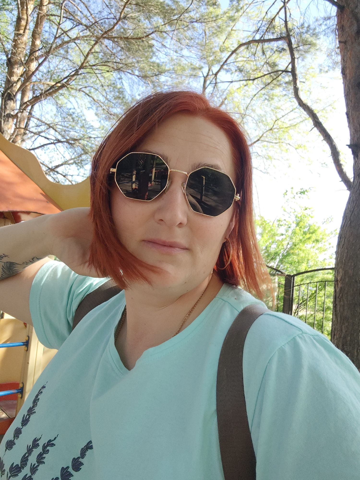 A woman with red hair is posing for a selfie outdoors. She is wearing sunglasses and a light blue t-shirt. A gold chain is visible around her neck. She is standing near a tree, with playground equipment visible in the background. The location is Chaikovsky, Russia.