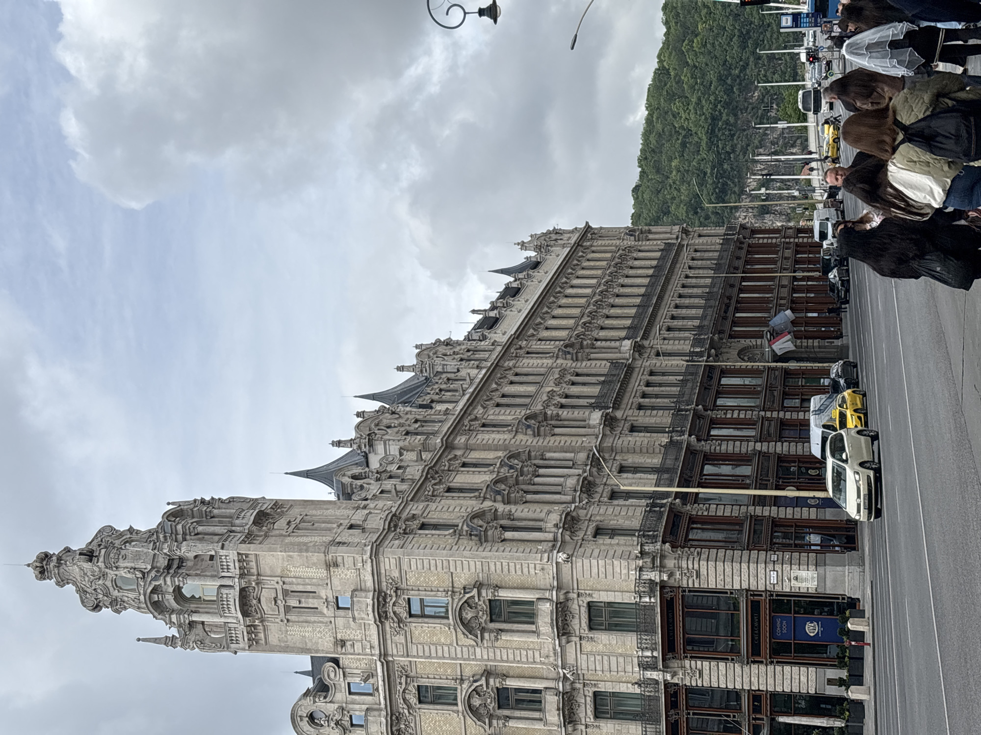The image depicts the Gresham Palace in Budapest, Hungary. The building is tall, ornate, and built in the Art Nouveau style. It faces a wide street with traffic, including cars and buses, and pedestrians walking on the sidewalk. On the opposite side of the street, a green, tree-covered hill is visible. The sky is overcast with grey clouds.
