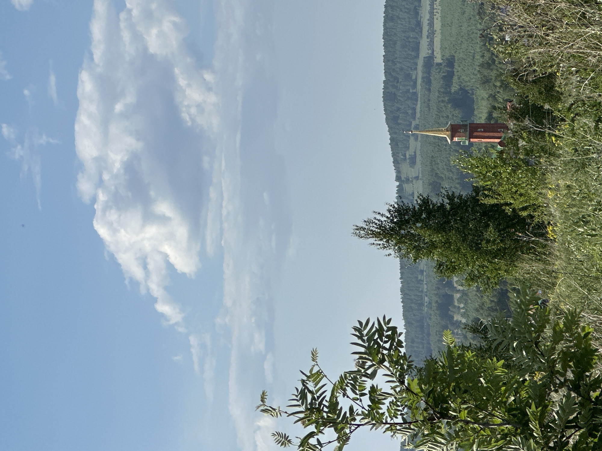 The image depicts a tower, possibly a church steeple, situated among trees, with a landscape of hills in the background. The sky is light blue with scattered white clouds. The foreground includes green foliage. The tower features a gold spire and a red structure. The image seems to be taken at Unknown, Unknown.