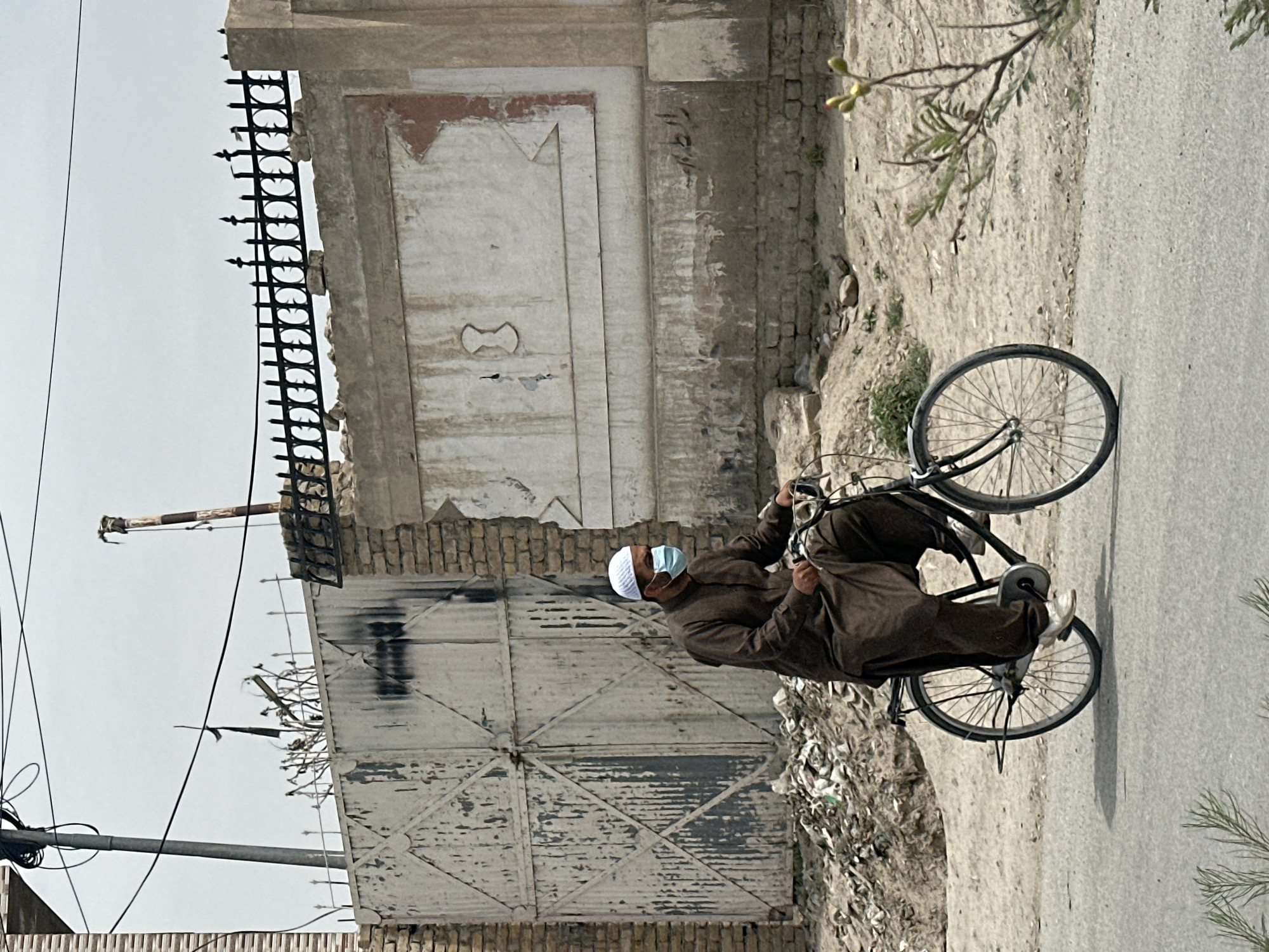 A man wearing a brown outfit, a white skullcap, and a light blue face mask is riding a black bicycle on a gray paved road. The man is facing to the right in the frame.
To the left of the man and bicycle is a weathered wall with a large, gated structure. The gate has a geometric pattern. Adjacent to the gate, further up the wall, is an ornate, rectangular panel with a lighter color. On top of the wall is a black, wrought-iron fence.
The environment appears to be an urban area, with a mix of aged buildings and some vegetation. Trash is visible next to the gate. Utility poles and wires are seen in the distance. The sky is overcast and light gray.