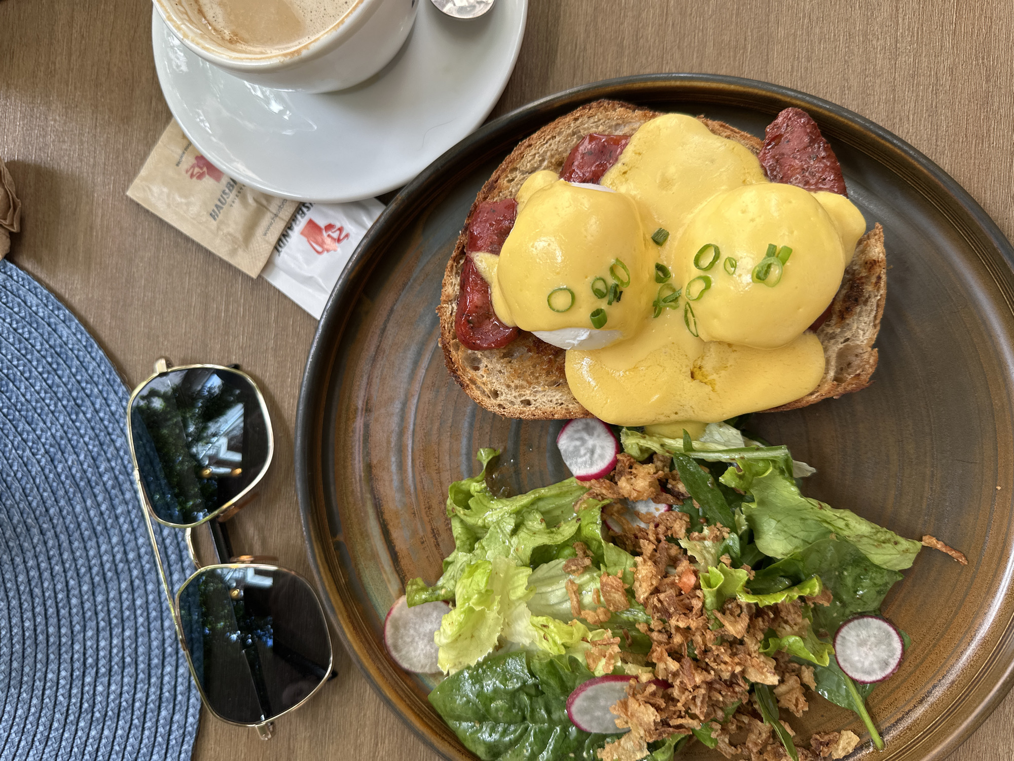 The image shows a close-up, overhead view of a breakfast meal on a wooden table. The meal, presented on a rustic, dark-rimmed plate, consists of Eggs Benedict served on toast, accompanied by a side salad. The Eggs Benedict features two poached eggs topped with a generous amount of hollandaise sauce and garnished with chopped green onions. Reddish-brown slices, likely sausage or bacon, are visible beneath the eggs. The salad is composed of fresh green lettuce leaves, sliced radishes, and topped with crispy fried onions.
In the background, a blue woven hat is positioned to the left of the plate, and a pair of aviator-style sunglasses with gold frames rests on the hat. To the right of the plate, a white teacup and saucer hold a coffee with a foamy top. Two small packets, one brown and one white, with red logos, are placed between the hat and the coffee cup. The overall setting suggests a casual outdoor dining experience, possibly at a cafe in Sofia, Bulgaria, given the context provided. The lighting is natural and bright, highlighting the textures and colors of the food and objects. No people are visible in the frame.