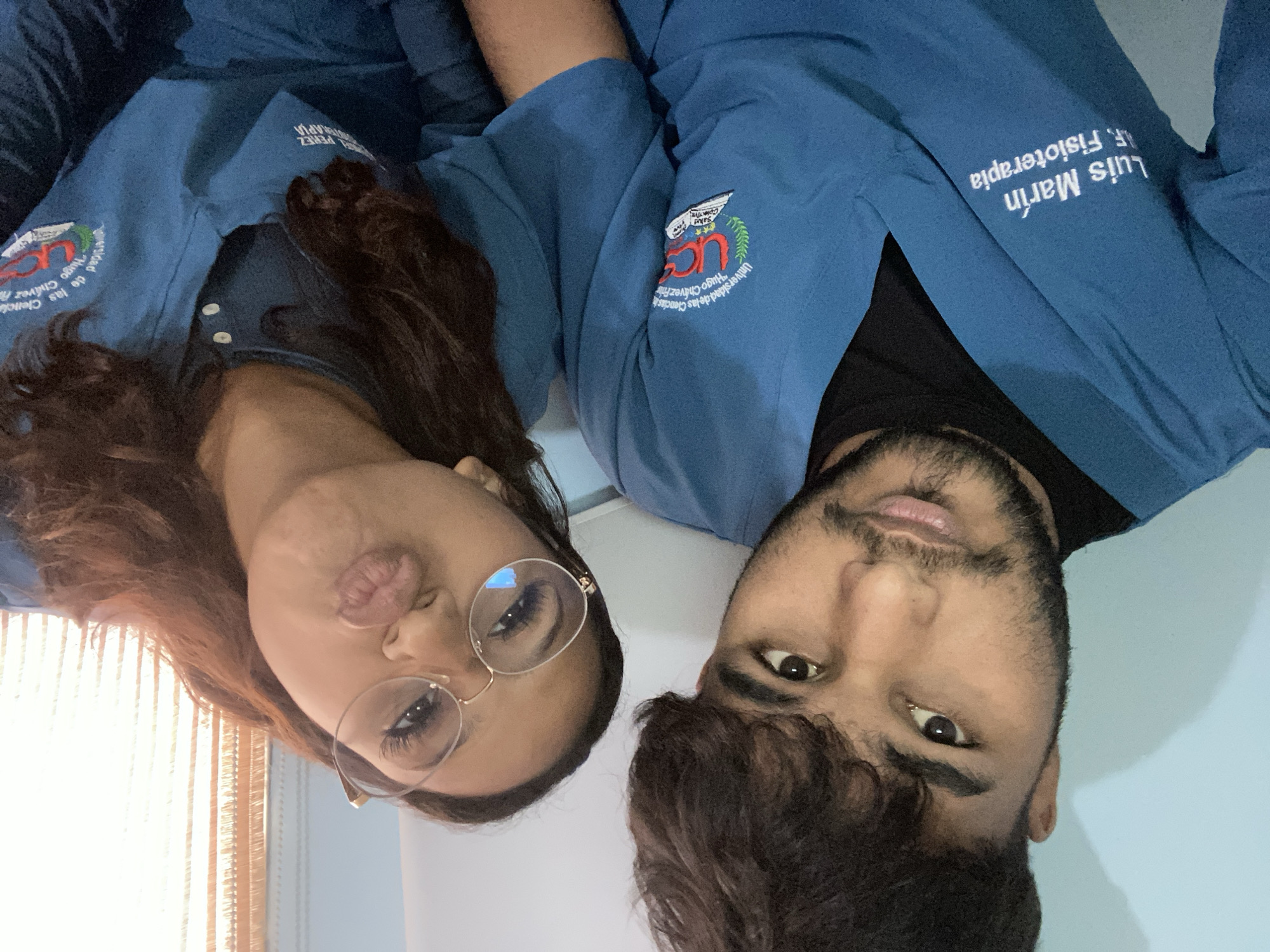 This media file is a close-up, rotated portrait of two young adults wearing blue scrubs, likely medical or healthcare professionals. The woman on the left has long, dark hair and is wearing glasses. She is puckering her lips, giving a playful expression. The man on the right has dark, curly hair and a beard. Both are looking towards the camera.
Visible text on the scrubs indicates they are affiliated with the "Universidad de las Ciencias Hugo Chávez Frías" and mention "Fisioterapia" (Physiotherapy). The woman's scrub has "Sakd PERÉZ TERAPIA" and a logo with "UCS". The man's scrub has "Luis Marín".
The lighting suggests an indoor setting, possibly near a window with blinds, given the light filtering from the left side of the image. The overall mood appears casual and friendly, suggesting they might be colleagues or friends. The background is a plain light blue wall. Given the location context of La Guardia, Venezuela, this could be a picture taken at their workplace or during a break.