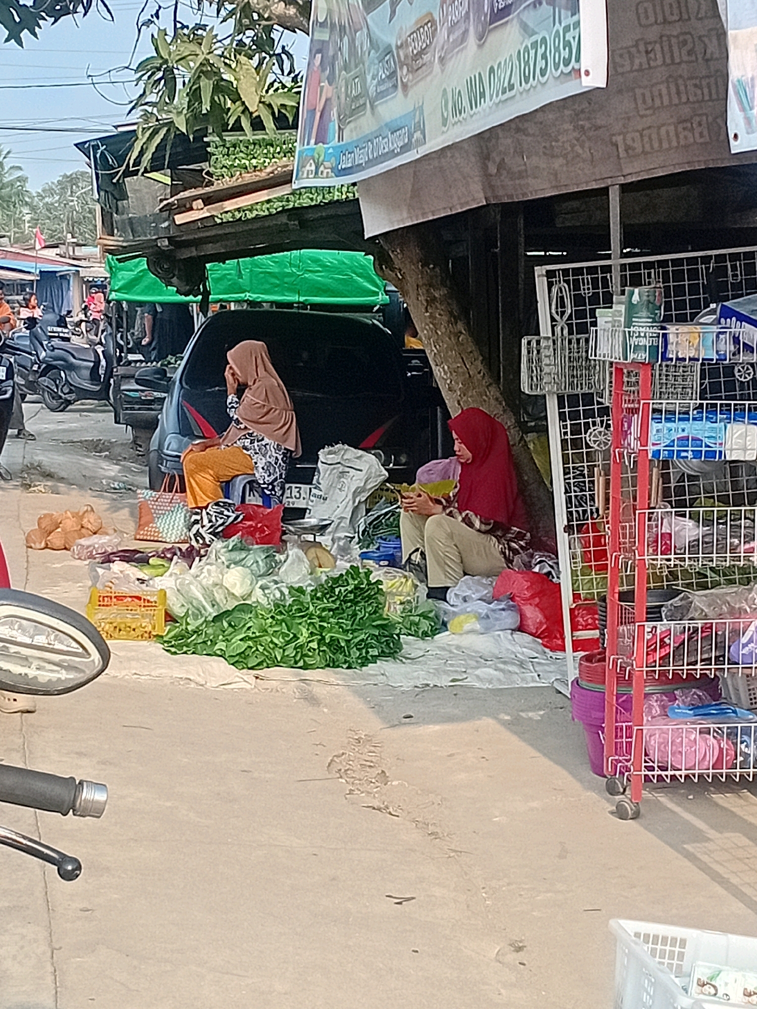 The image depicts a street-level scene in Anggana, Indonesia, likely a marketplace. In the foreground, the handlebars and part of the front wheel of a motorcycle are visible. A woman wearing a light brown hijab and patterned shirt is seated on a small stool on the left side of the image, facing away from the camera, with her right hand touching her face. In front of her are several shopping bags and a pile of produce, including leafy greens and what appear to be onions or similar vegetables. To the right of this woman, a second woman, wearing a red hijab and beige pants, is seated on the ground, facing left, and appears to be looking at a yellow object in her hands. Behind the women, a dark-colored car is parked, partially obscuring a stall or structure. A red display rack filled with various items, including plastic containers and packaged goods, is on the far right of the image. The ground is paved and appears somewhat dusty. The background shows more stalls and greenery, suggesting a vibrant commercial area.