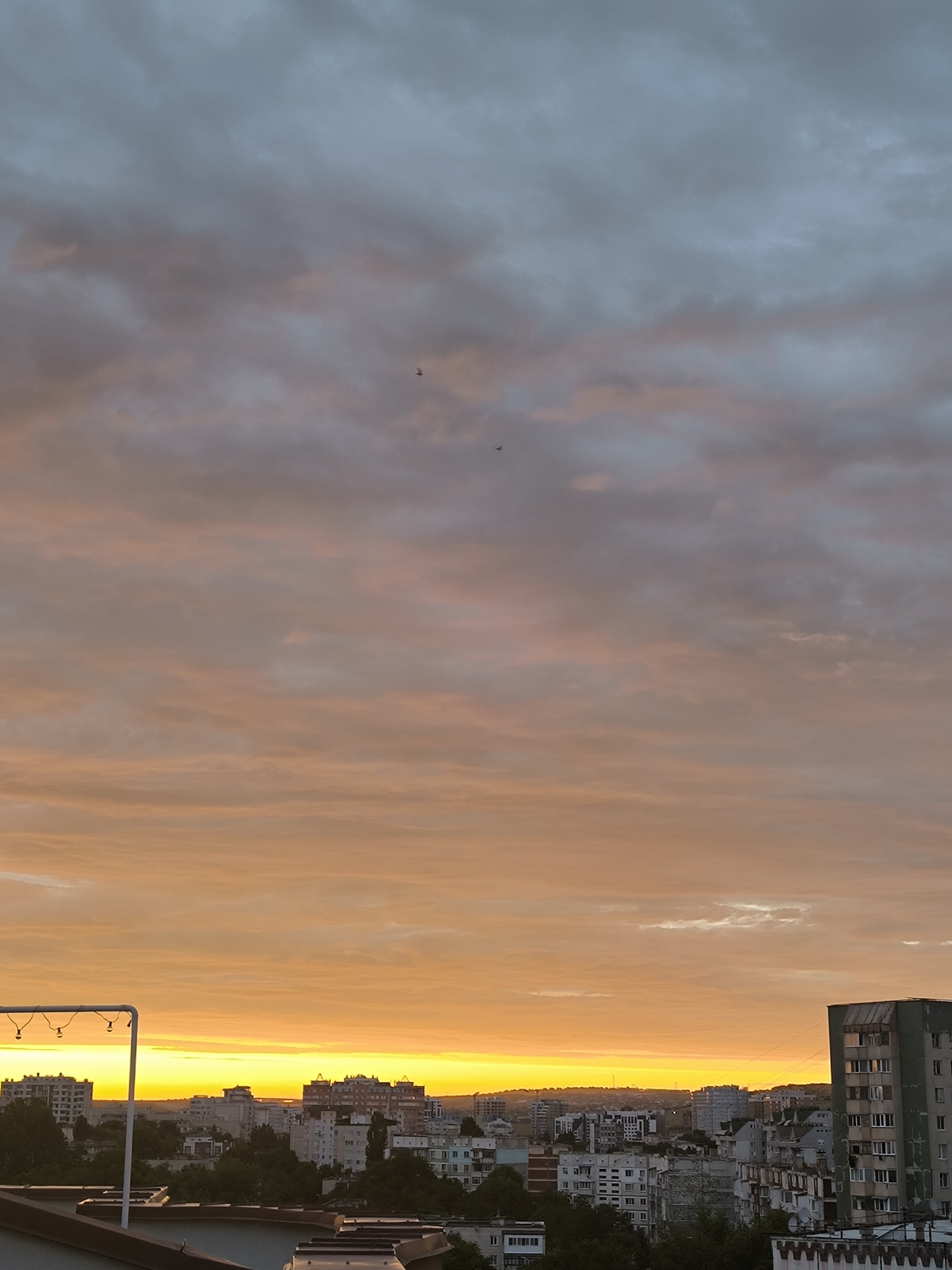 This is a vertical, eye-level, outdoor, medium shot photograph of a city skyline during sunset. The sky takes up the majority of the frame, with a gradient of colors from bright yellow and orange at the horizon to darker gray and blue at the top. The clouds are textured and appear to be illuminated by the setting sun. Two small, dark shapes, possibly birds or airplanes, are visible in the sky.
Below the sky is a dense urban landscape, likely a residential area, with numerous buildings of varying heights and architectural styles. Many of the buildings are apartment blocks, some appearing older with faded paint and weathered facades. There are also some trees interspersed among the buildings. The foreground shows the roof of a building, with what appears to be outdoor lighting fixtures attached to a white structure. The overall impression is that of a city preparing for evening under a dramatic sunset.
Based on the architectural style and the presence of specific building types, this scene is consistent with a view of the city of Chișinău, Moldova. The specific location appears to be from an elevated vantage point, possibly a rooftop, overlooking a neighborhood such as Telecentru or Botanica. The time of day is sunset, as indicated by the vibrant colors in the sky and the low angle of the sun. There are no visible people in the frame.