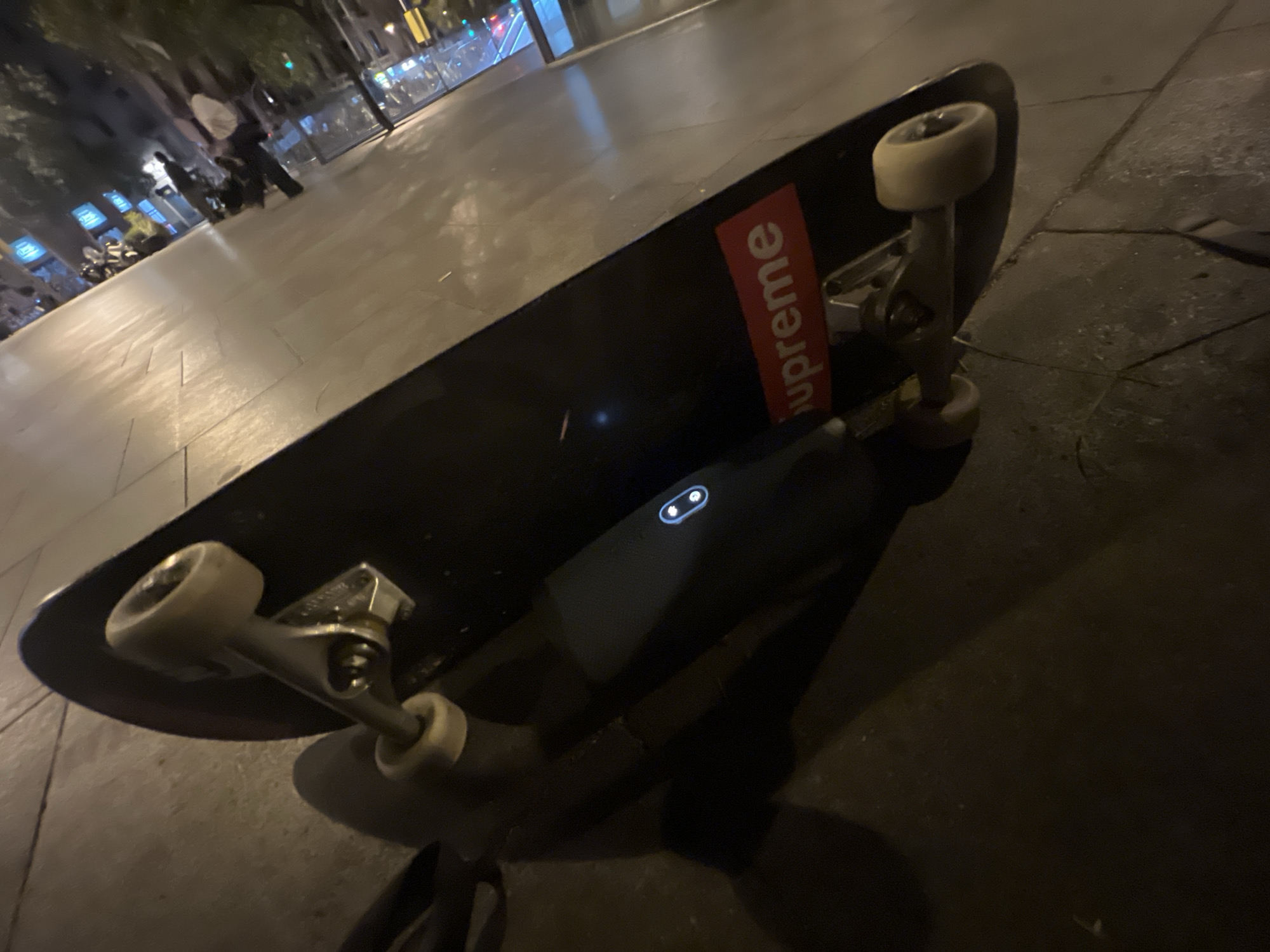 The image is a close-up, low-angle shot of a black skateboard with white wheels, resting on a paved surface at night. A prominent red "Supreme" sticker is affixed to the underside of the skateboard deck. The skateboard is angled diagonally across the frame. In the background, slightly out of focus, appear to be urban elements: streetlights, some building facades with illuminated windows, and a few indistinct figures. The paving stones suggest an outdoor public space, consistent with a city sidewalk or plaza. The lighting conditions indicate it is dark outside, with artificial light sources illuminating the scene. The overall impression is of a skateboard left unattended in a city environment at night.