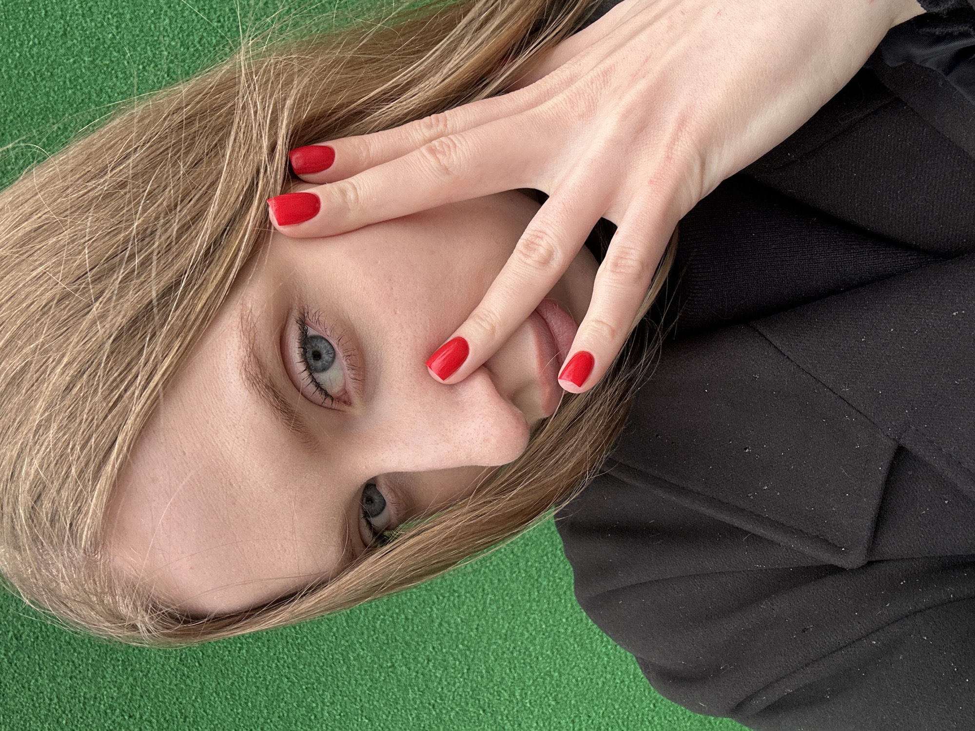 An eye-level close-up shot captures a young woman with fair skin and light brown hair, posing with her left hand partially covering her face. The image is tilted, giving a dynamic feel. Her left hand, adorned with bright, matte red nail polish on each visible fingernail, is spread across her cheek and mouth, with fingers slightly parted.
One of her light blue eyes is clearly visible, looking directly towards the viewer, framed by dark eyelashes. Her skin appears smooth with some visible pores, and strands of her straight hair fall across her forehead and temple. She is wearing a black garment, likely a coat or jacket, made of a textured fabric that shows some fine dust or fibers, especially on the shoulder area.
The background consists of a uniform, textured green surface, resembling artificial turf or a vibrant green carpet, which contrasts with her fair skin and dark clothing. There is no visible text in the image. The pose suggests a moment of shyness, playfulness, or a deliberate artistic composition. The setting, given the location context of Chaikovsky, Russia, could be an indoor space or an outdoor area with artificial green flooring.