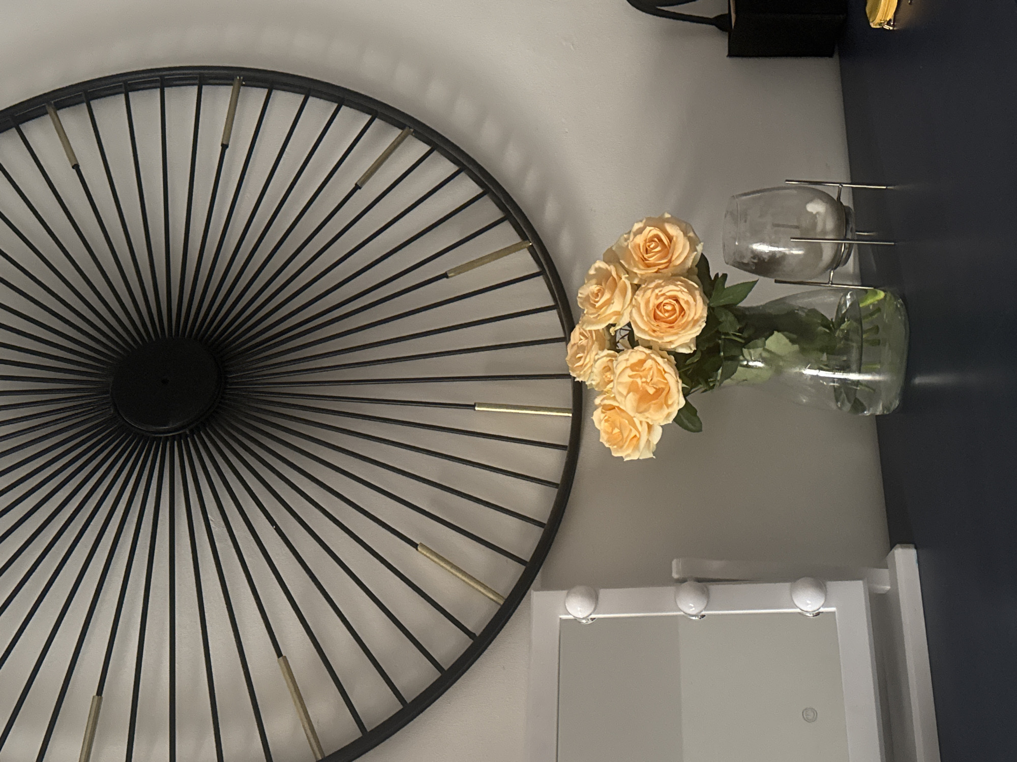 A close-up shot shows a decorative black metal sunburst design mounted on a light gray wall, with metallic gold accents at intervals along its rays. To the right, a clear glass vase holds a bouquet of peach-colored roses, positioned near a white-framed vanity mirror with round bulbs. Above these elements, a corner of a dark blue surface is visible, with a small decorative object and a clear glass on a metal stand. There is no evidence to support the location of this image being in the city of Bat Yam, Israel.