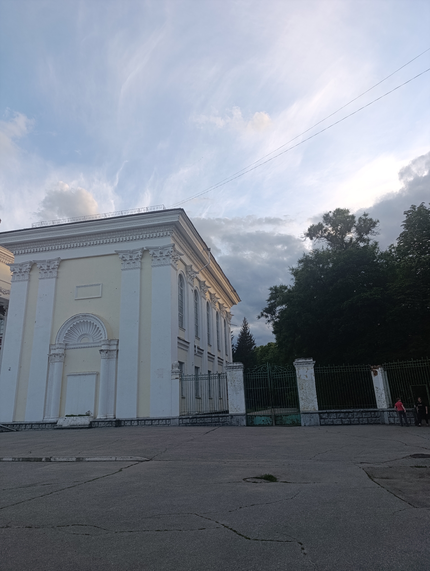 The image shows the exterior of a pale yellow building with white classical architectural elements, including columns and decorative molding. The building is partially visible, with its corner facing the viewer. A green metal fence with white pillars runs along the front of the building, separating it from a paved area. Tall, dark green trees are visible behind the fence. The sky above is a mix of pale blue with wispy white clouds and darker, more ominous clouds in the upper right quadrant. Two people are standing near the fence on the right side of the image; one is wearing a red shirt and the other is wearing dark clothing. The paved area in the foreground is cracked and appears to be a pathway or street.