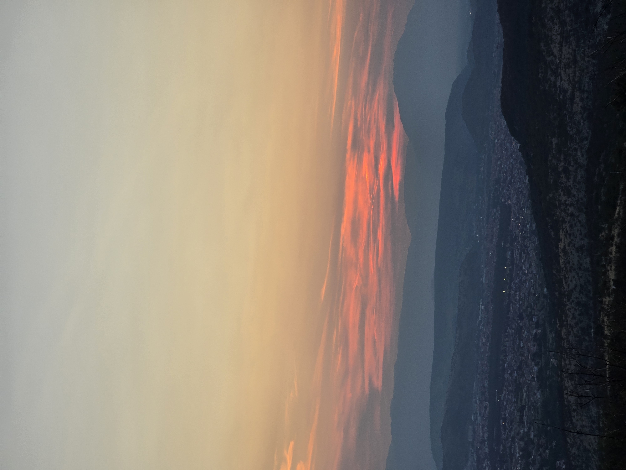 The image depicts a panoramic view of Donja Vrbica, Montenegro, at dusk. The sky transitions from a pale yellow at the top to a vibrant orange and pink band of clouds near the horizon, suggesting the setting sun. Layered mountain ranges fade into the distance, becoming progressively darker and more silhouetted against the sky. In the midground, a densely populated town with visible streetlights is nestled within a valley. The foreground is dominated by dark, silhouetted hills and sparse vegetation, including bare branches of trees. The overall atmosphere is serene and natural, with the urban landscape softly illuminated by evening lights.