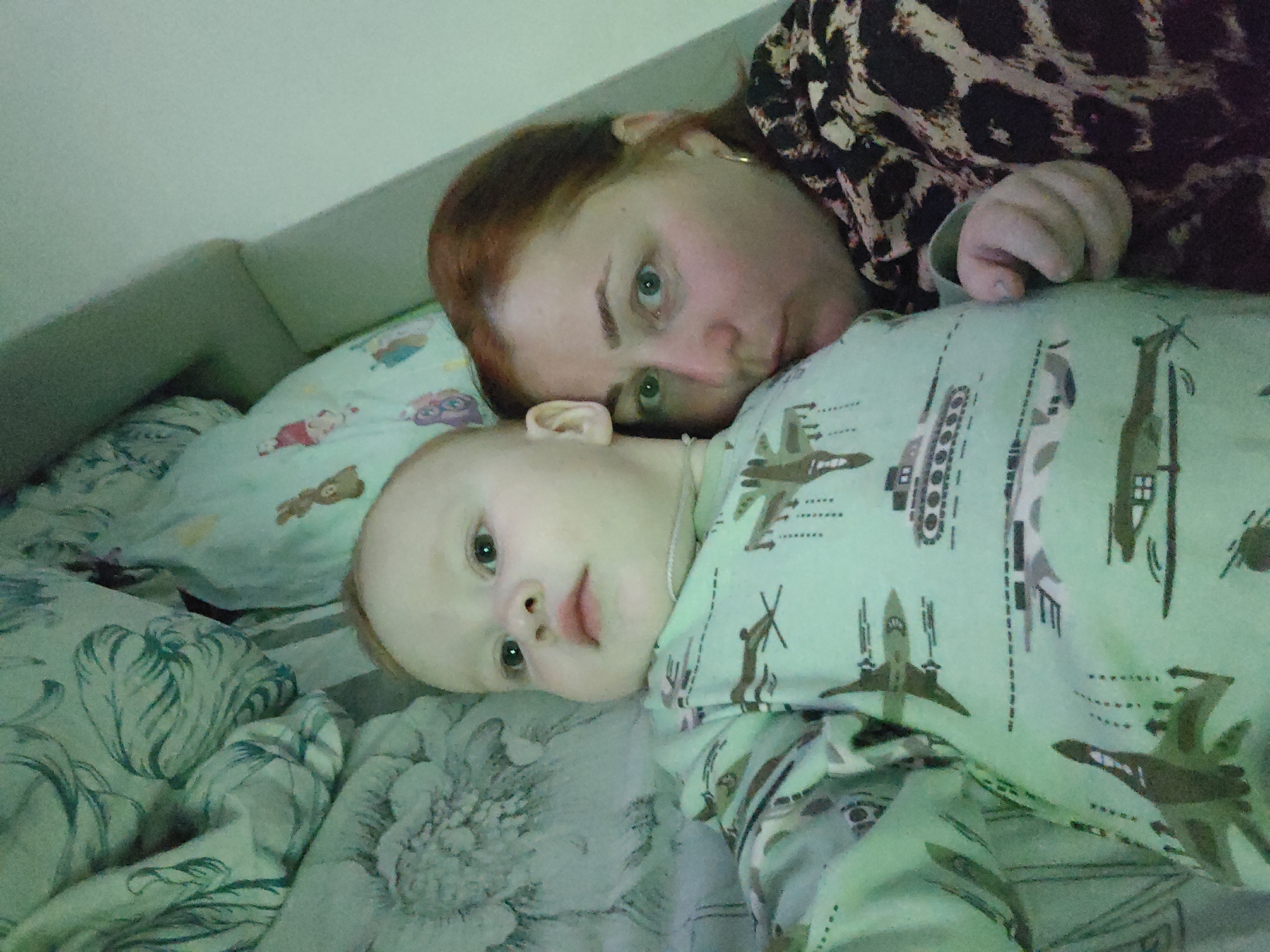A woman with red hair and green eyes lies in bed with a baby dressed in pajamas with a military vehicle pattern. The background includes bedding with a floral pattern. The picture appears to be taken in an indoor setting. No specific details indicate the location being Markovskii, Russia.