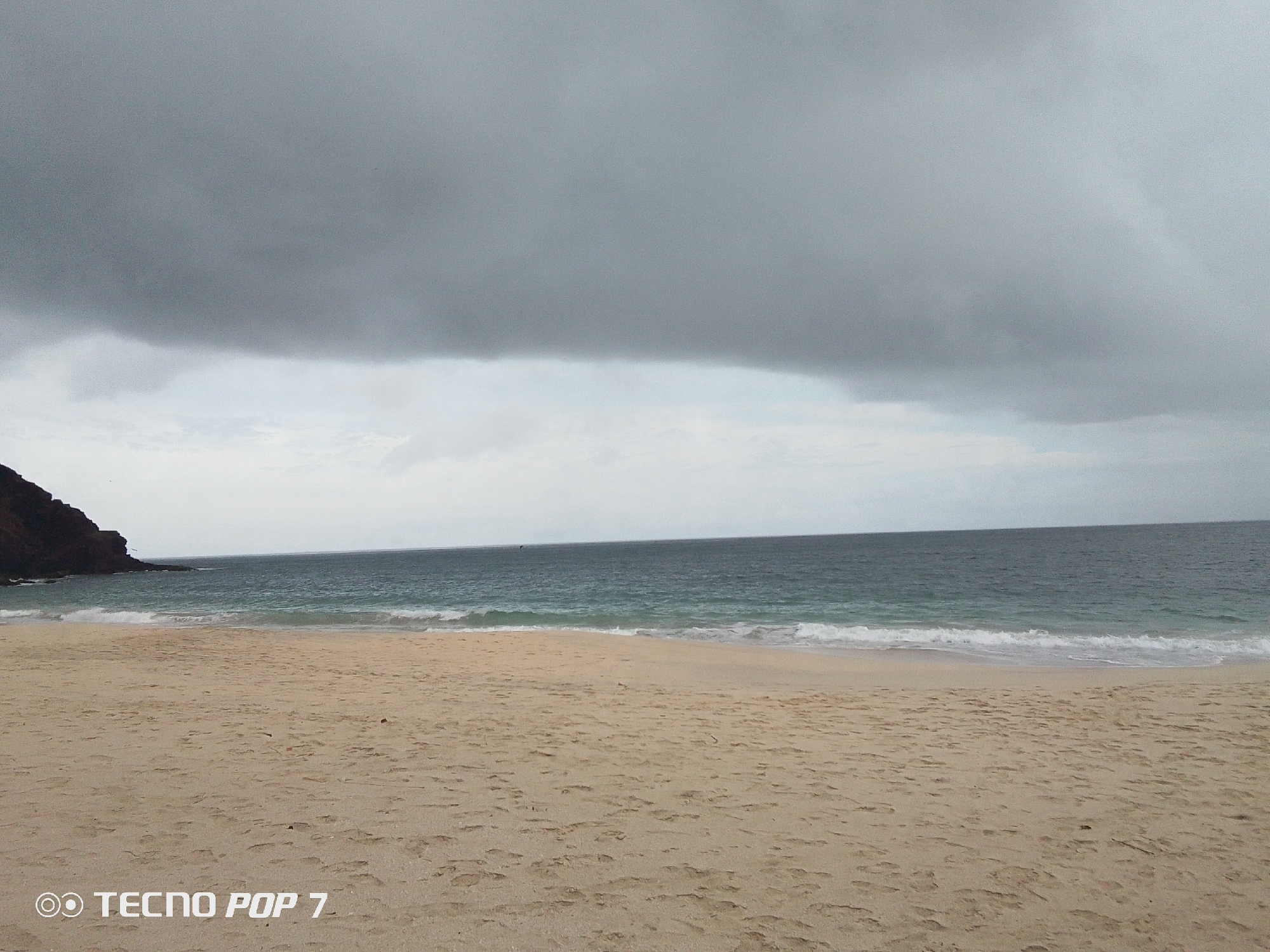 The image depicts a beach scene under a heavily overcast sky in Pedro González, Venezuela. A small, rocky outcrop is visible on the left. The sand is tan, and the water is a dark blue-gray. Gentle waves are washing onto the shore. The "TECNO POP 7" watermark is located at the bottom left. The scene appears deserted with no people visible.