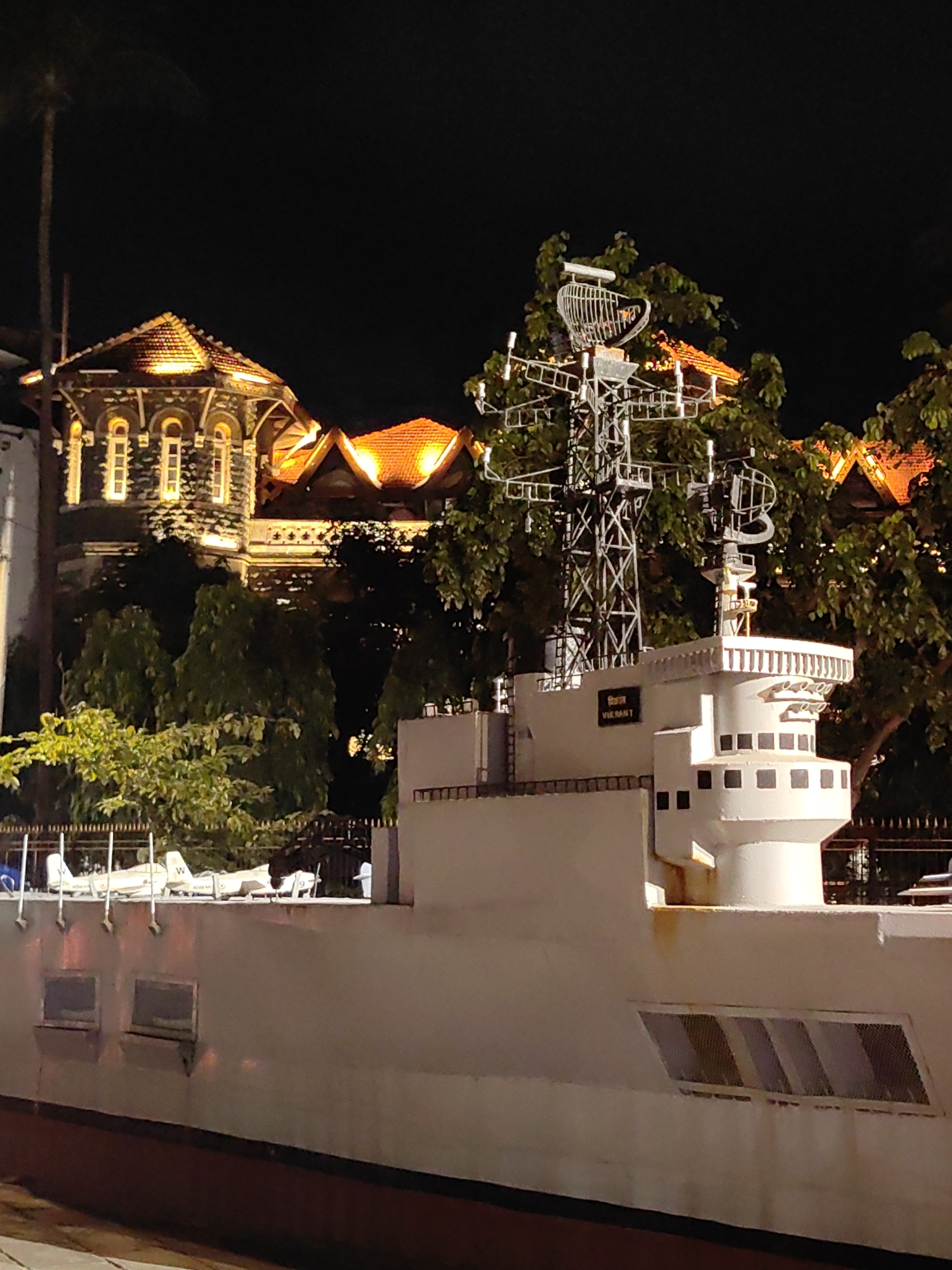 The image shows a nighttime view of a model of the INS Vikrant, a former aircraft carrier of the Indian Navy, at the Indian Naval Maritime Museum in Mumbai, India. The model is positioned in the foreground, with its white hull and superstructure clearly visible. A prominent radar mast rises from the deck. In the background, a building with ornate architecture and brightly lit tiled roofs is illuminated, with trees visible between the building and the model ship. The sky is dark, indicating it is night. The museum is located at the Gateway of India in Colaba, Mumbai.