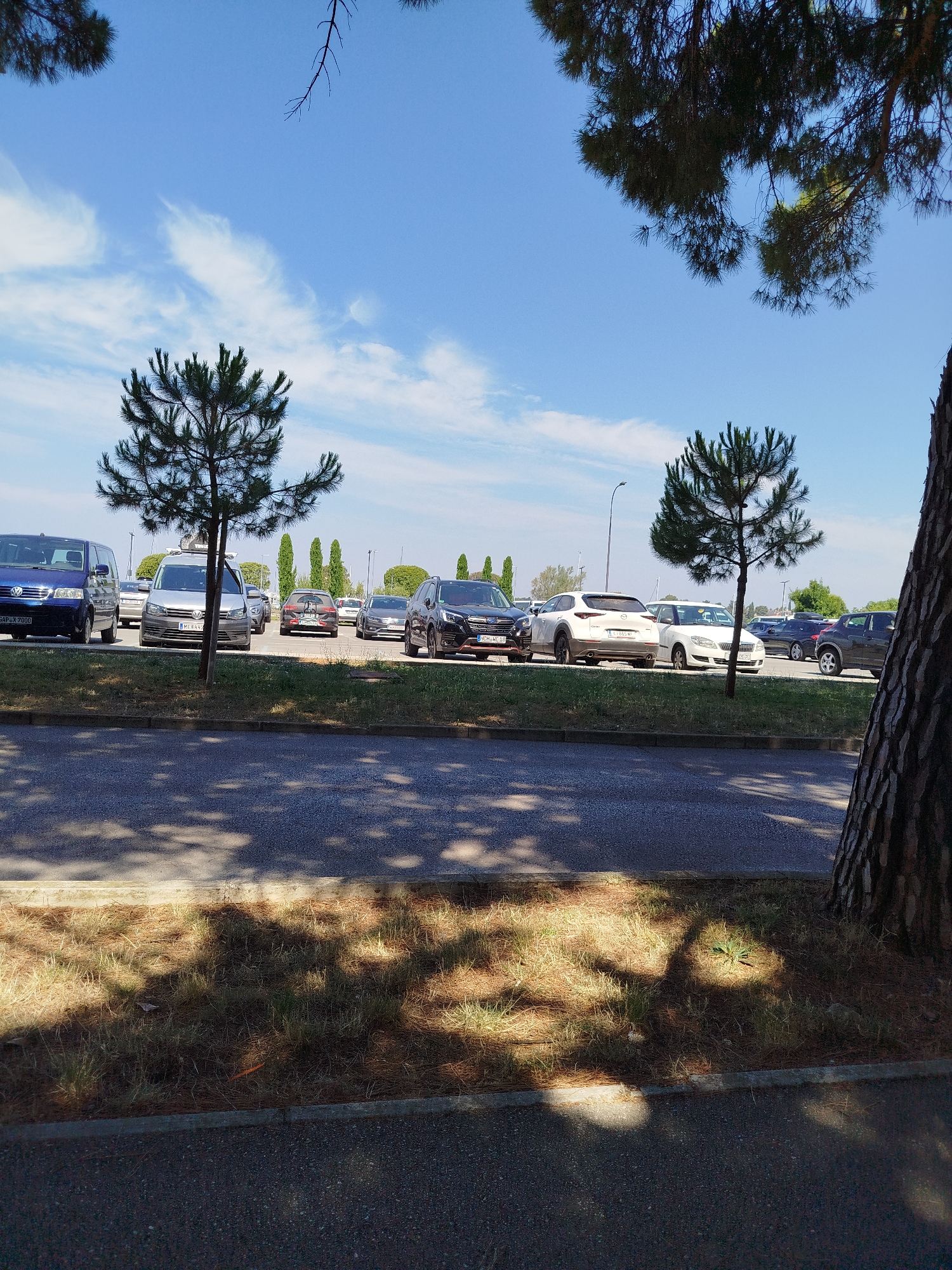 This image depicts an outdoor parking lot on a sunny day, likely in Umag, Croatia. The scene is dominated by parked cars, with a few thin, evergreen trees scattered throughout the lot. In the foreground, a paved area and a grassy strip with scattered pine needles are visible, casting dappled shadows from overhead trees. The sky is blue with scattered white clouds, suggesting fair weather.
Several cars are visible in the parking lot, including a blue minivan on the left, a silver van, a black SUV, and a white hatchback. Some of the cars have license plates, though the text is not clearly legible in most cases. One license plate on the blue van appears to read "SAPX 7000". Another on the silver van shows "ME 644WZ". A white car has a license plate with the text "CH 682 WI". A dark SUV has a license plate with "WCH...10". The parking lot seems to be situated near a body of water, possibly the sea, as evidenced by distant masts of sailboats. The overall impression is of a typical day in a parking area, likely during daylight hours given the bright sunshine and clear shadows. No people are visible in the image. The atmosphere is serene and uneventful.
