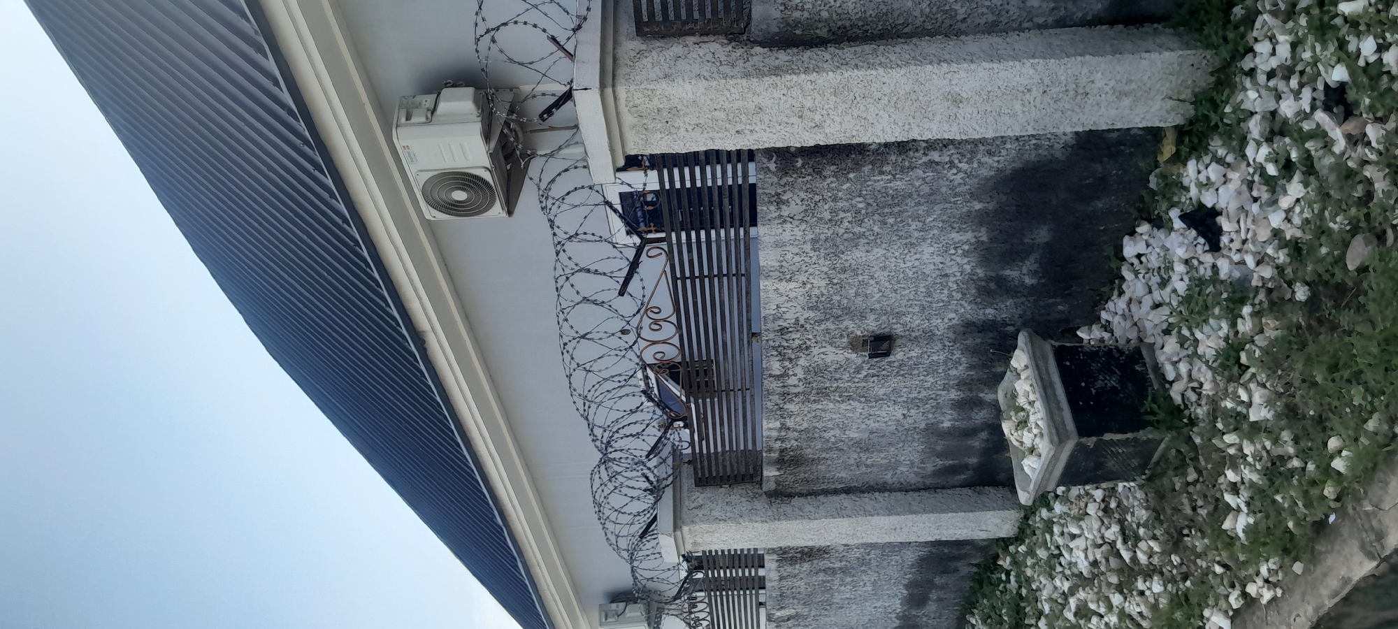 The image depicts a residential property in Jalingo, Nigeria, characterized by a light gray building with a dark corrugated metal roof. A prominent feature is the security fencing, consisting of a concrete wall topped with horizontal metal bars and a coiled barbed wire entanglement along the top. Two outdoor air conditioning units are mounted on the exterior wall of the building. In the foreground, a concrete planter box filled with light-colored stones and some sparse vegetation sits on a ground covered with similar stones and green grass. The scene is set against a clear blue sky. There are no people visible in the image.