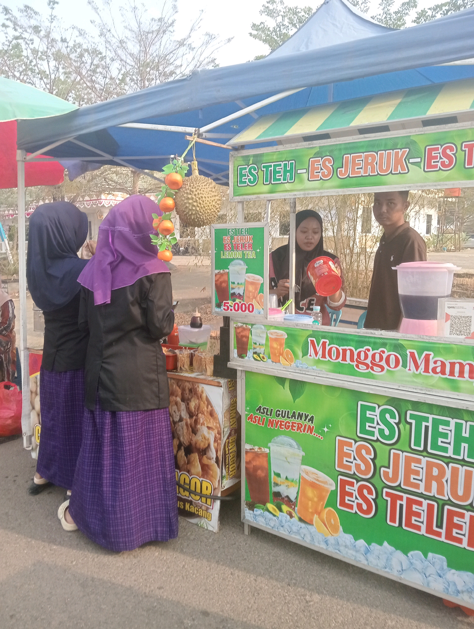 In Sidoharjo, Indonesia, a street vendor stall displays various iced drinks and snacks. The stall is covered by a blue and green awning with a yellow and green striped border. In the foreground, a white and green sign advertises "ES TEH - ES JERUK - ES TELER" with images of drinks. To the left, two individuals wearing hijabs and long skirts are seen from behind. One is wearing a dark blue hijab and the other a purple one. Behind them, a durian fruit hangs from a string, alongside decorative orange fruits. Further back, a woman in a pink headscarf and a man in a brown shirt are attending the stall. The woman is holding a red container, and a pink drink dispenser is visible. To the right of the women in hijabs, a sign with an image of fried food, possibly "gorengan" or "bakso goreng," is displayed, with text indicating "US Kacang." The pavement is a light gray asphalt.