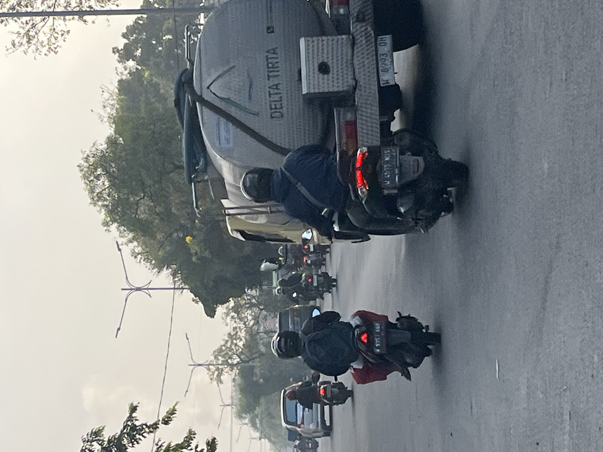 This image captures a busy street scene in Gelam, Indonesia. A silver water tanker truck with the logo "DELTA TIRTA" prominently displayed on its side is traveling down the road. Several individuals are riding motorcycles, their forms partially obscured by helmets and casual clothing. The environment appears to be an urban setting with streetlights and trees lining the road, indicating it's likely daytime with overcast skies. The presence of multiple vehicles and riders suggests a typical traffic flow in the area.
