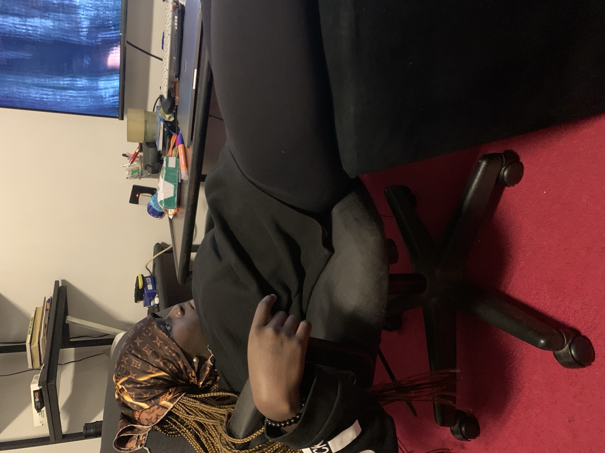 A woman, with braided blonde hair and wearing a brown and gold patterned headscarf, is shown from the side in a reclined position in a black office chair on a red carpet. She is wearing a black top and black leggings. Her left hand is resting on her lap, and she is looking to her right. In the background, there is a desk with various items including books, pens, a laptop, and a television screen displaying a blue, textured image. A black shelving unit is also visible.