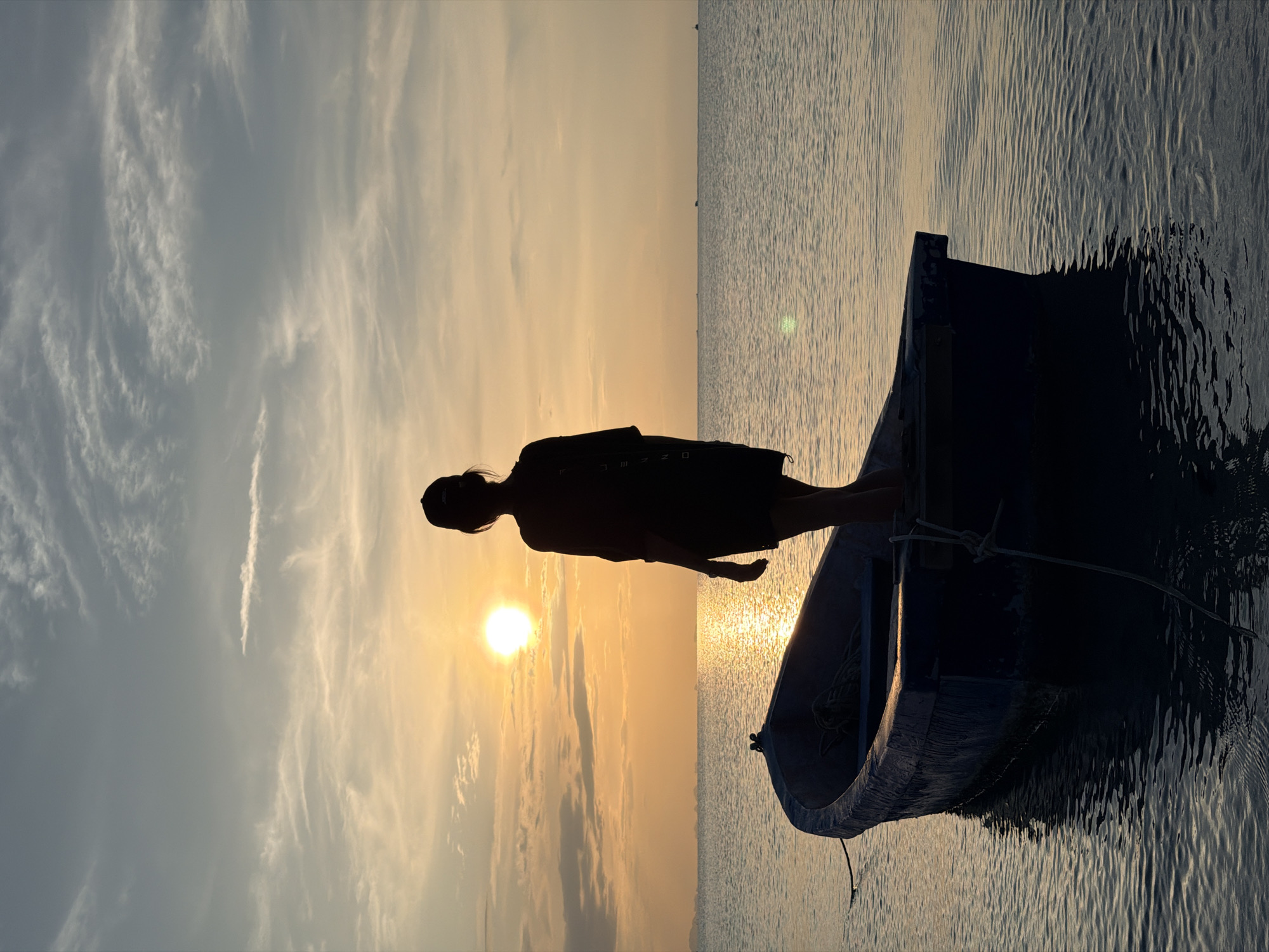 A full-frame, vertical shot captures a silhouetted person standing in a small blue boat on calm water. The person, wearing a dark shirt and shorts and a cap, faces away from the camera, looking towards a bright, setting sun. The sun is positioned in the upper left quadrant of the frame, casting a golden glow across the sky and reflecting on the water. The sky is filled with scattered clouds, with hues of orange and yellow near the horizon, transitioning to blue and gray higher up. The boat, with a rope tied to its side, is partially submerged in the water. The overall scene is tranquil and evokes a sense of solitude. The location is identified as Koh Samui District, Thailand.