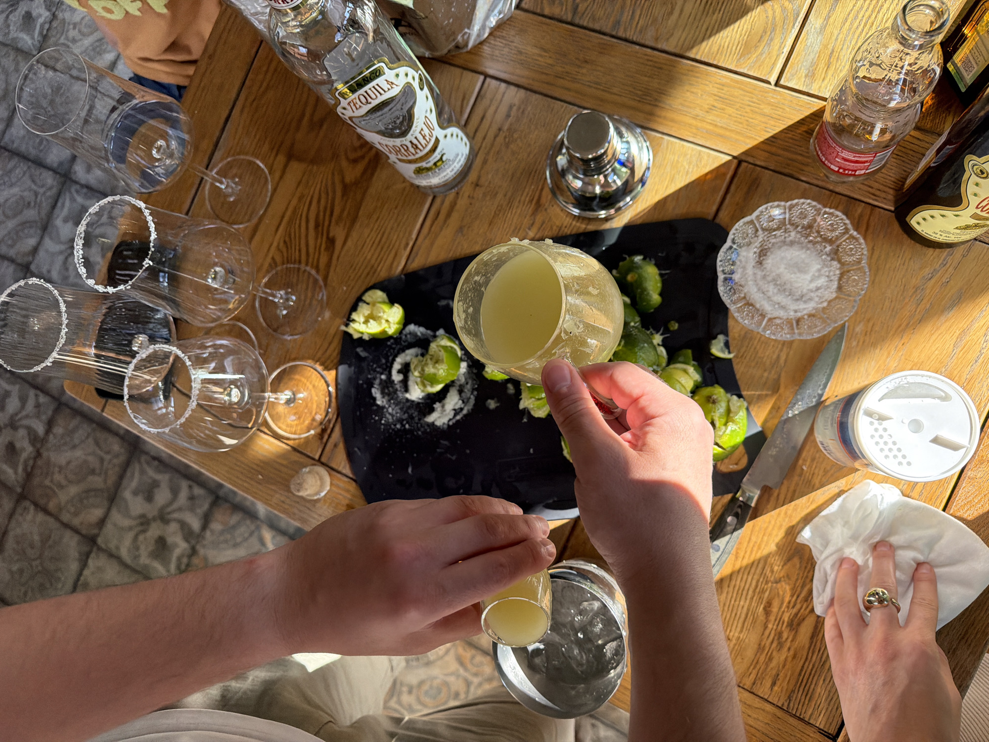 This image depicts a top-down view of a table outdoors, possibly in Mishutkino, Russia, as suggested by the context provided. The scene is set on a wooden table, with some tiled flooring visible in the background. Several clear glasses with salt-rimmed edges are arranged on the table. A bottle of "Corralejo Blanco Tequila" is lying on its side, with a small portion of its label visible. There are also several cut lime wedges scattered on a black cutting board.
Two individuals' hands are visible. One hand is pouring a pale yellow liquid from a small shot glass into a larger glass containing ice. The other hand is holding a white napkin and appears to be preparing to wipe something. A silver knife with a black handle is lying next to the cutting board. There is also a small, decorative glass dish filled with salt, and a small silver shaker. In the foreground, another hand is visible wearing a ring. Further down the table, another bottle, partially obscured, is present. The sunlight casts distinct shadows across the table, indicating it's daytime.