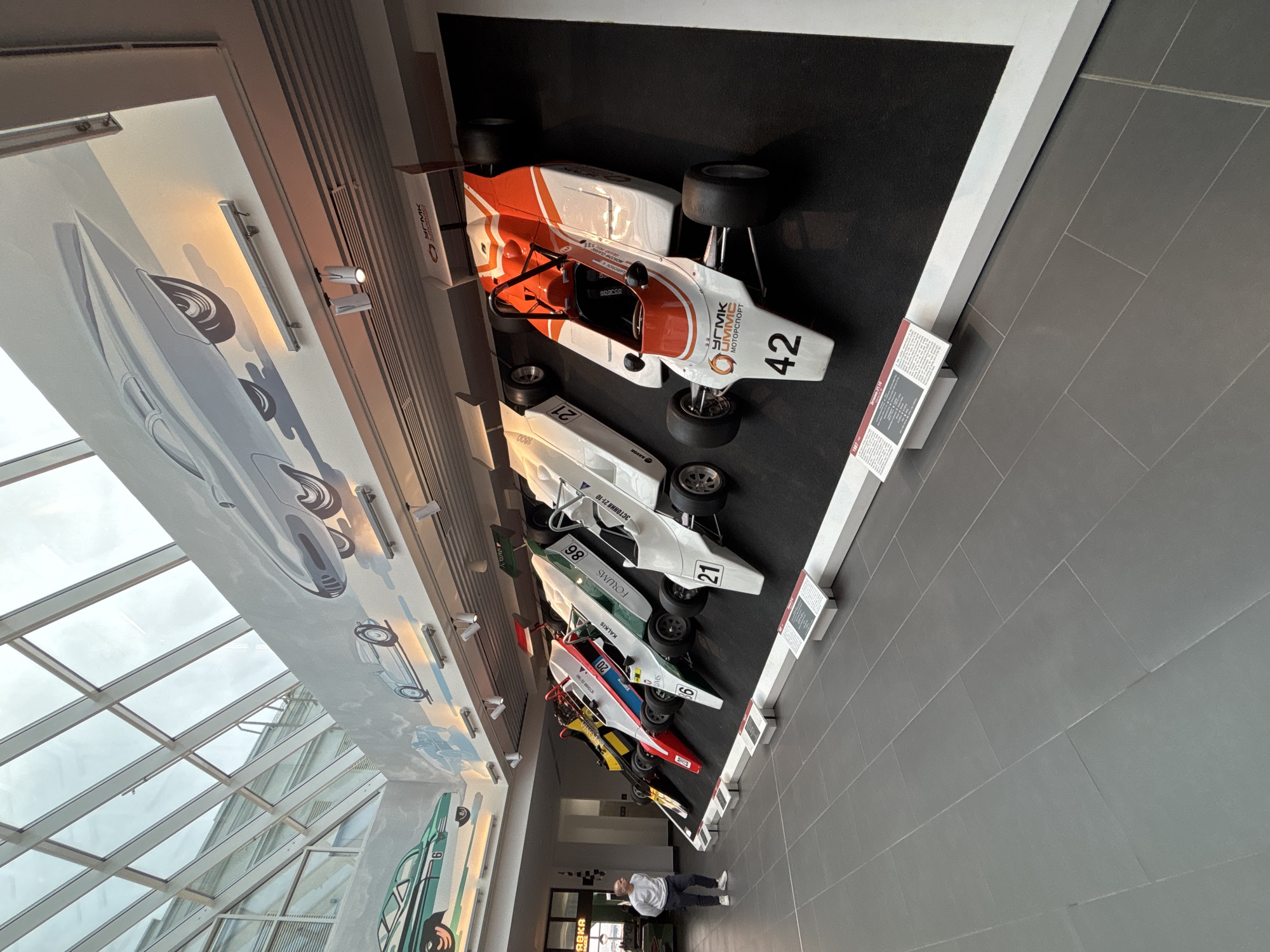 This image depicts a museum exhibit featuring a row of vintage Formula 1-style race cars displayed on a black elevated platform. The cars are white with various colored accents and numbers. The largest and most prominent car in the foreground is white and orange with the number 42. Behind it are several other cars, including one with the number 21, another with 86, and one with 20. A man in a grey sweater and dark pants stands to the left, observing the exhibit. The background features a large mural of stylized cars and a sloped glass ceiling. Information plaques are placed in front of the cars on the floor. This exhibit is located in the UMMC Automotive Museum in Verkhnyaya Pyshma, Russia, which is a suburb of Yekaterinburg.