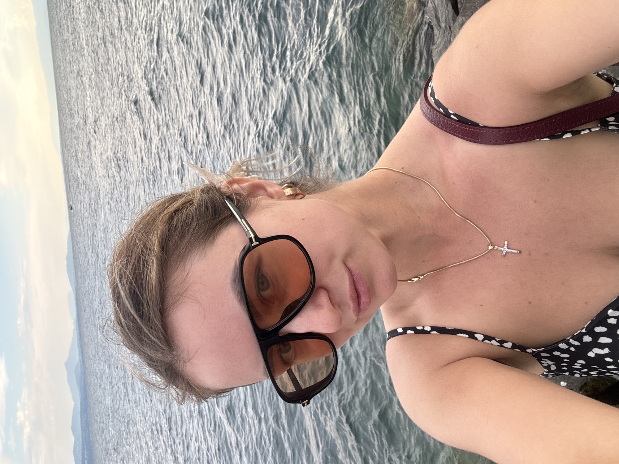 A woman with blonde hair pulled back, wearing large black sunglasses with amber lenses, a gold necklace with a cross pendant, and a black and white polka dot top, takes a selfie outdoors. She is standing on a rocky shore with the calm, rippling water of Lake Geneva stretching out behind her. In the distance, the hazy outlines of mountains are visible against a pale sky. The scene is illuminated by soft, natural light, suggesting it is either early morning or late afternoon. The woman's expression is neutral. She appears to be at a waterfront location in Lausanne, Switzerland, likely enjoying a view of the lake and the surrounding landscape.