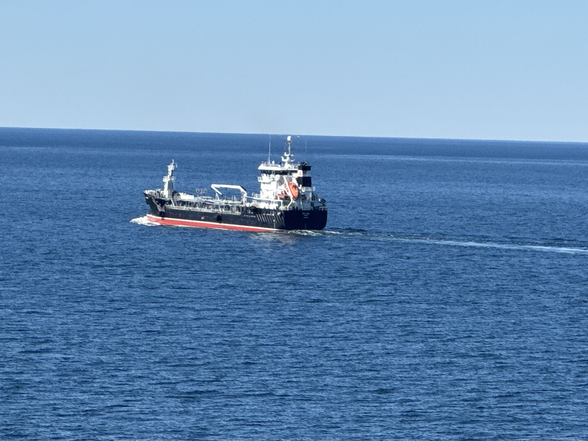 A modern cargo ship, dark hull with a red waterline and white superstructure, sails through calm blue ocean waters under a clear sky. The ship is observed from a slightly elevated perspective, showing its port side. The vessel is leaving a white wake trailing behind it. The horizon line is distinct, separating the water from the pale blue sky. The ship is heading towards the right of the frame. This scene is likely offshore, not within the immediate city limits of Karlskrona, Sweden, but potentially in the waters surrounding the archipelago.