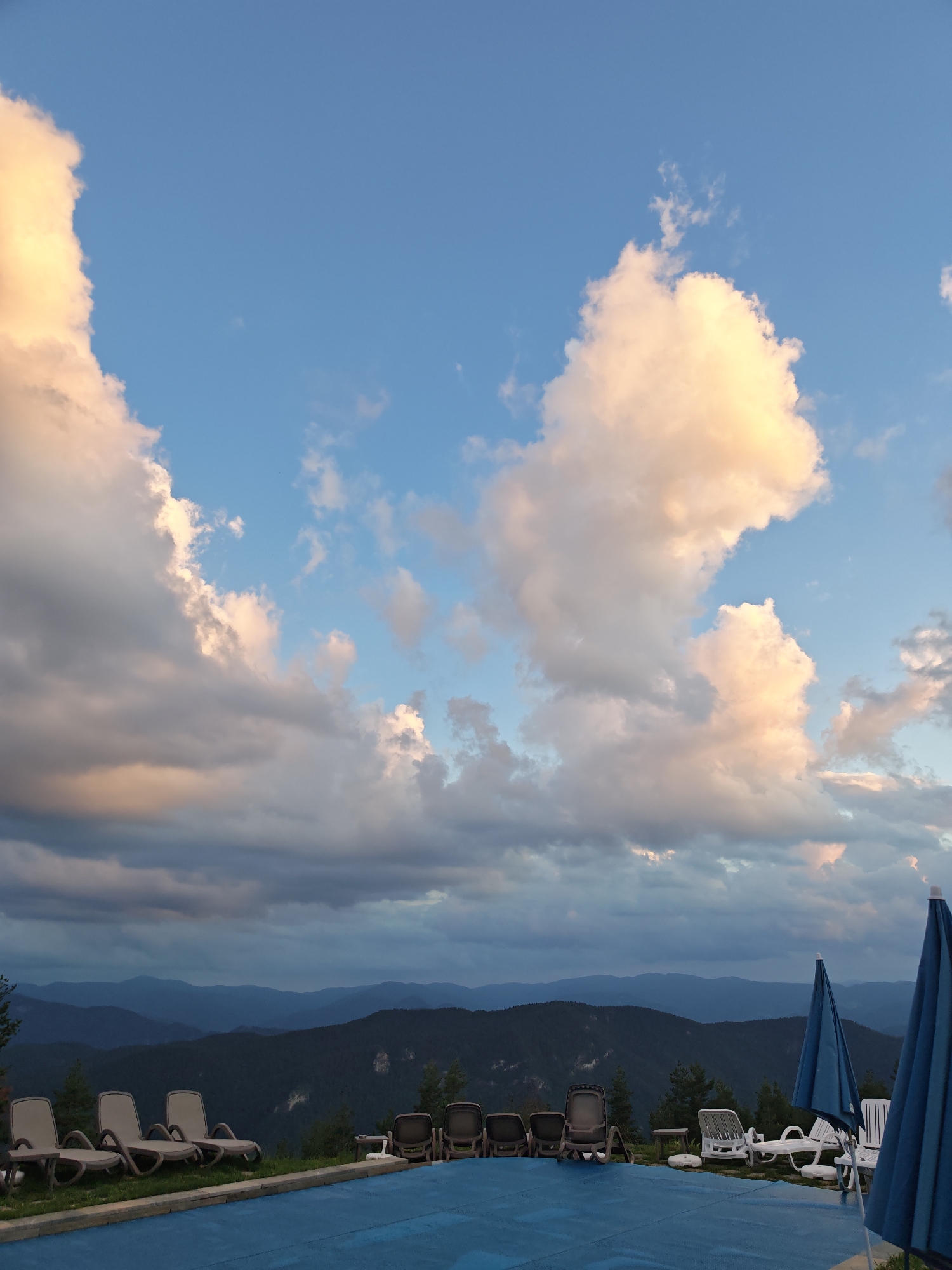 The image captures a scenic view of a swimming pool area overlooking a mountain range, likely in the vicinity of Dobrostan, Bulgaria. The sky is dominated by dramatic clouds, with hues of orange and yellow suggesting either sunrise or sunset. Several lounge chairs are arranged around the pool, some empty and others occupied by a few individuals. Two large blue umbrellas are also visible, providing shade. The pool itself is covered with a blue tarp. The background consists of layered, dark green mountains receding into the distance under the cloudy sky.