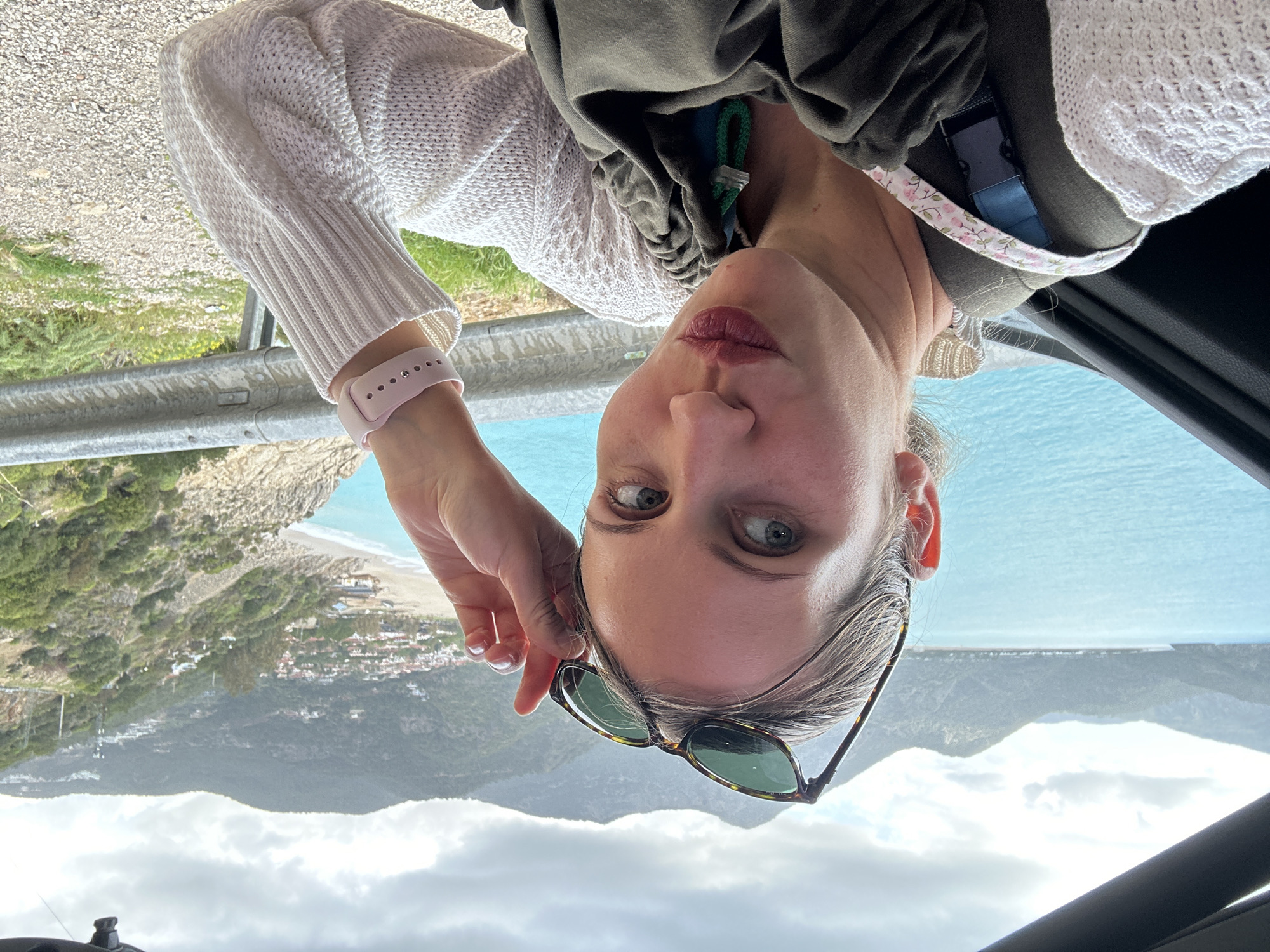 A selfie of a woman with gray hair, wearing a white knit sweater and a pink smart watch, is oriented upside down. She is holding a pair of sunglasses in her right hand. Behind her is a coastal landscape. The mountains are visible in the background, along with a view of the Mediterranean Sea. The scene is possibly taken near Fethiye, Turkey, due to the appearance of the coastal environment and mountain range. The bottom of the image includes the interior of a car.