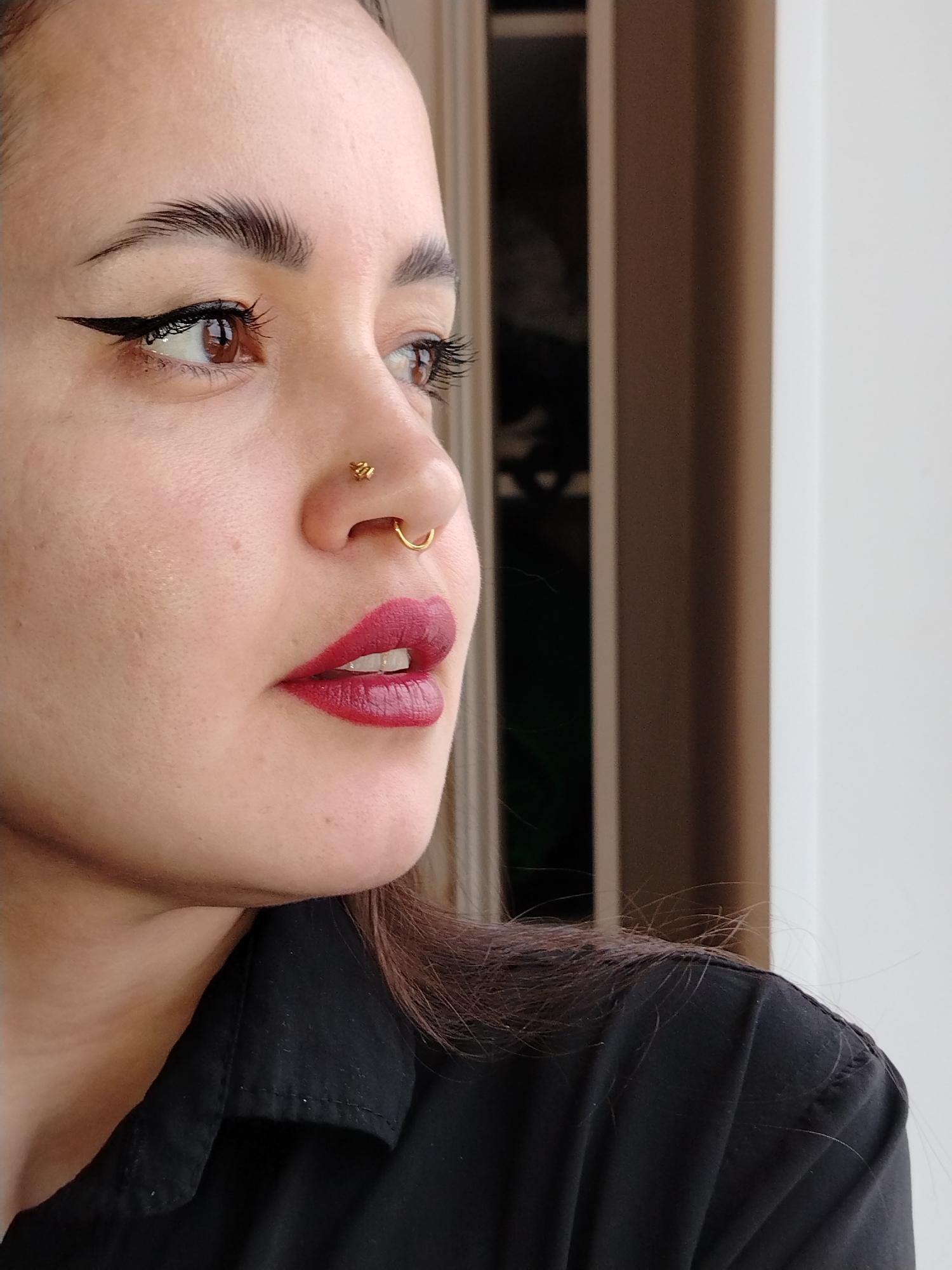 A close-up shot depicts a woman with fair skin, brown eyes, and dark hair, looking to the side, likely towards a window. She has dark eyeliner, well-defined eyebrows, dark red lipstick, and gold nose rings. She is wearing a black button-down shirt. The background includes a door frame and a white wall. The location is unknown.