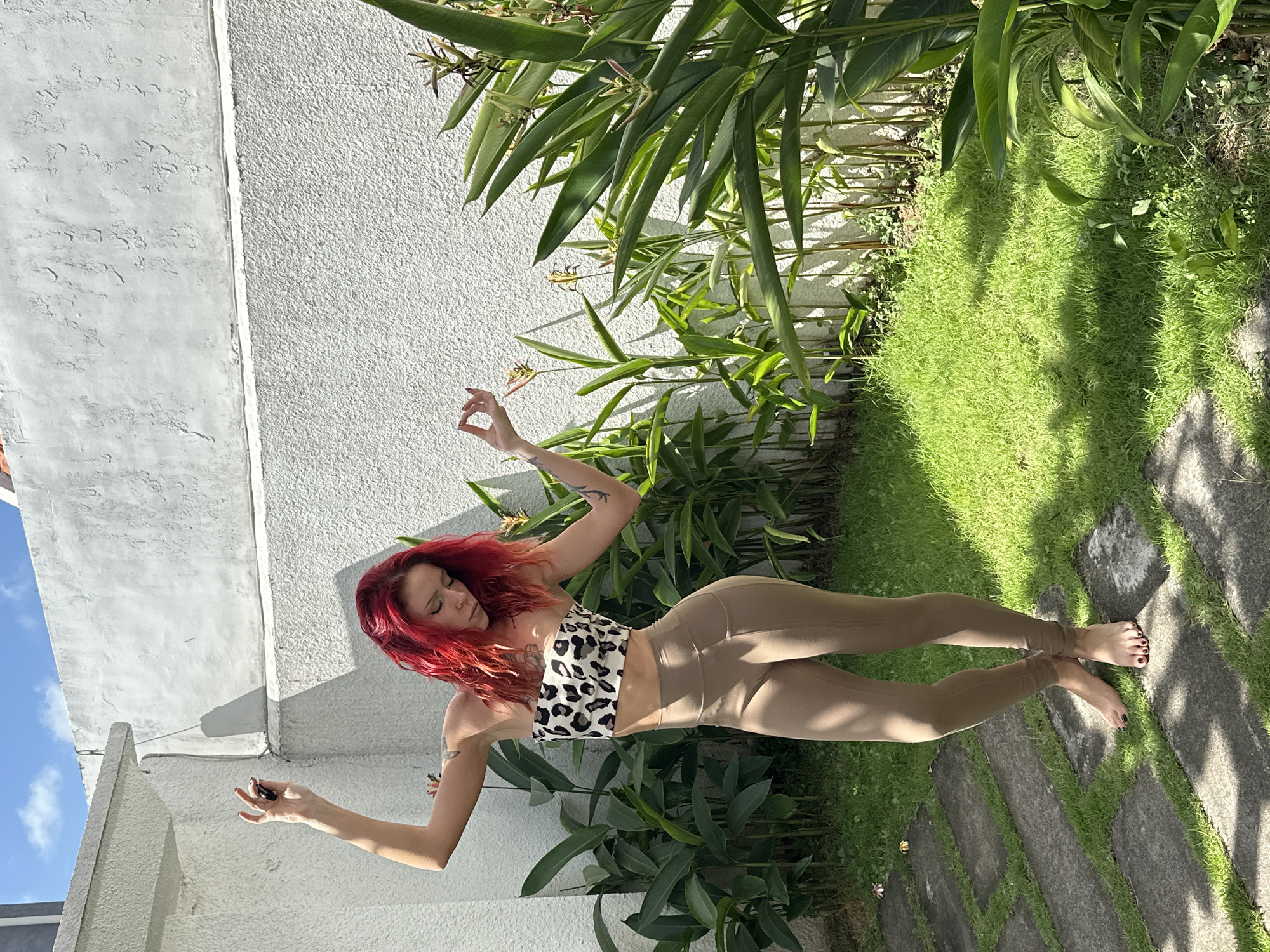A woman with vibrant red hair is captured in a full-body shot, outdoors in what appears to be a garden setting. She is wearing a leopard print bandeau top and tan leggings. Her body is angled, with her arms extended outwards in a pose that suggests balance or a dance-like movement. She is looking downwards with her eyes closed, conveying a sense of calm or focus.
The background features a textured white wall on the left side, with a section of blue sky visible above. To the right of the woman, lush green foliage and grass are prominent, with dappled sunlight casting shadows on the ground. Large stepping stones are laid out across the grassy area, forming a pathway. The scene evokes a peaceful, possibly meditative atmosphere. The time of day appears to be sunny, with clear skies and bright daylight. The location context indicates North Kuta, Indonesia. There is no visible text in the image.
