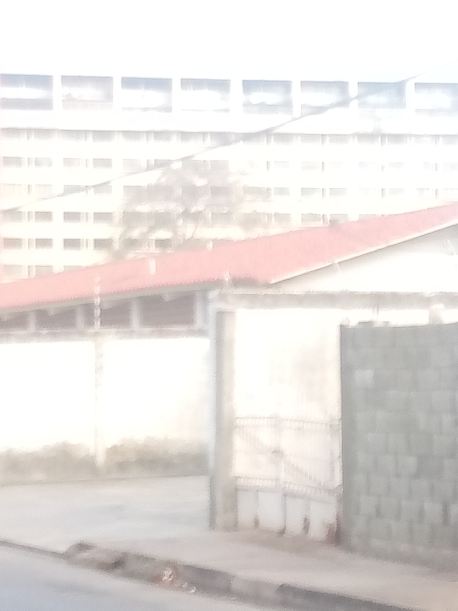 The image depicts an outdoor scene of a building with a red-tiled roof in the foreground. Behind this structure, a large, multi-story building with numerous windows is visible, appearing somewhat out of focus. The area in front of the red-roofed building is partially enclosed by a wall and a gate. To the right of the gate, a section of a brick wall is present. The ground in the foreground shows a portion of a street or sidewalk with a curb. The overall impression is of an urban or semi-urban setting with architectural structures.