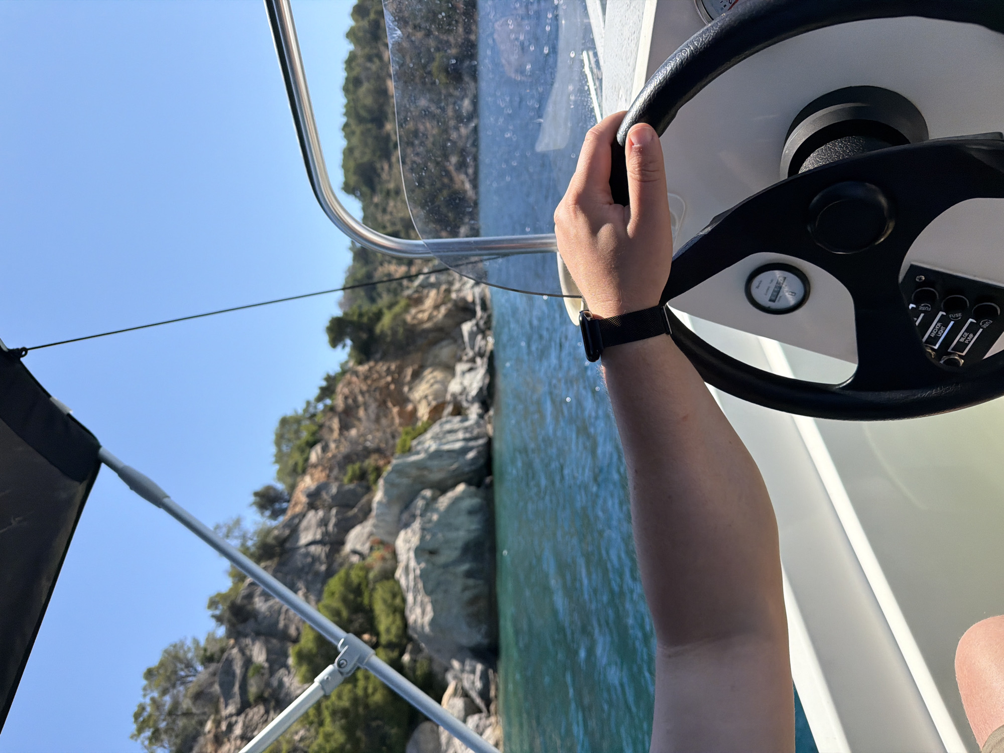 This image provides a first-person perspective from a boat navigating the waters around Poros, Greece. The foreground is dominated by a person's right hand gripping the black steering wheel of the boat. Their left forearm, adorned with a black smartwatch or fitness tracker with a mesh band, is also visible.
The boat's white dashboard is partially seen, featuring a circular gauge with numbers like '0', '40', '80', and '120', and a panel of toggle switches. Visible labels on these switches include "FUSE" (repeated), "ANCHOR LIGHT", and "BILGE PUMP". A clear plastic windshield separates the boat's cockpit from the open environment, with metal railings and supports for a canopy or bimini top visible on the left.
Beyond the boat, the sea stretches out, displaying clear, blue-green water that hints at shallow depths or excellent clarity. The background reveals a picturesque, rugged coastline. This shore is characterized by large, light-colored rocks and cliffs, interspersed with lush green trees and vegetation. The sky above is a brilliant, cloudless blue, indicative of a bright, sunny day, likely during the morning or afternoon. The scene evokes a sense of calm and leisure, suggesting a pleasant boating excursion along the scenic Greek coast.