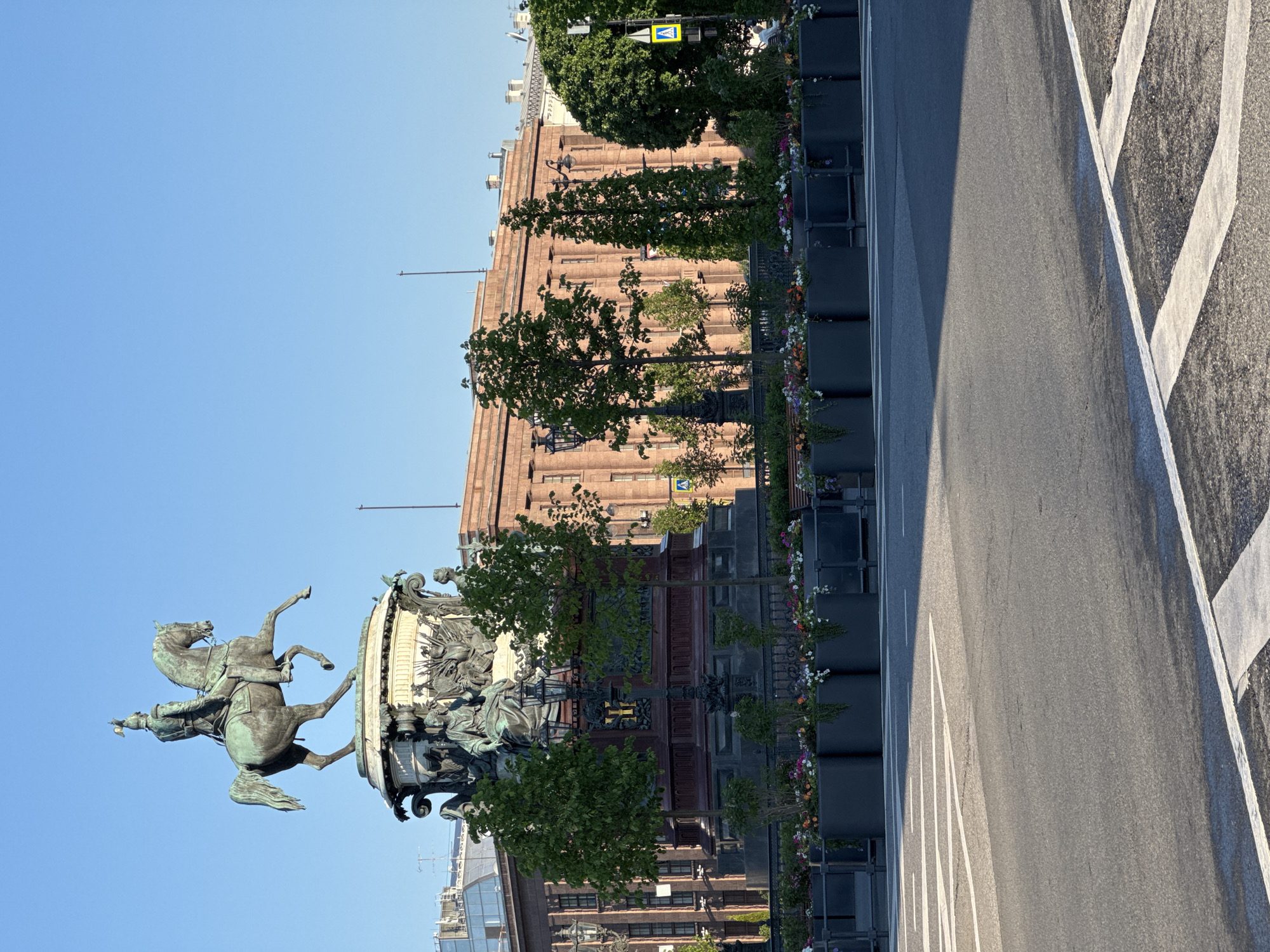 The image displays a sunny urban landscape in Saint Petersburg, Russia, centered around a prominent equestrian statue. In the foreground and left side, a grand bronze equestrian statue, likely the Monument to Peter the Great (the Bronze Horseman), captures attention. It features a figure on a rearing horse, its front hooves lifted, appearing to be balanced on a serpent. The statue rests upon a massive, irregularly shaped natural stone pedestal, embellished with intricate sculptural details at its base.
Moving towards the right, a row of green trees with full foliage lines the street, interspersed with dark, rectangular planters bursting with colorful flowers in shades of purple, white, orange, and pink. Behind this verdant line, a large, stately building with a reddish-brown facade and numerous windows provides a historical architectural backdrop.
An asphalt road occupies the midground and right side of the frame, marked with crisp white painted lines, including sections that suggest a pedestrian crossing. Above the trees on the right, a yellow diamond-shaped road sign with a black pedestrian symbol is clearly visible, indicating a crossing.
The bright, even illumination and clear shadows suggest a sunny late morning or early afternoon. The sky is a uniform, clear blue. The overall scene evokes the historical grandeur and vibrant public spaces characteristic of Saint Petersburg.