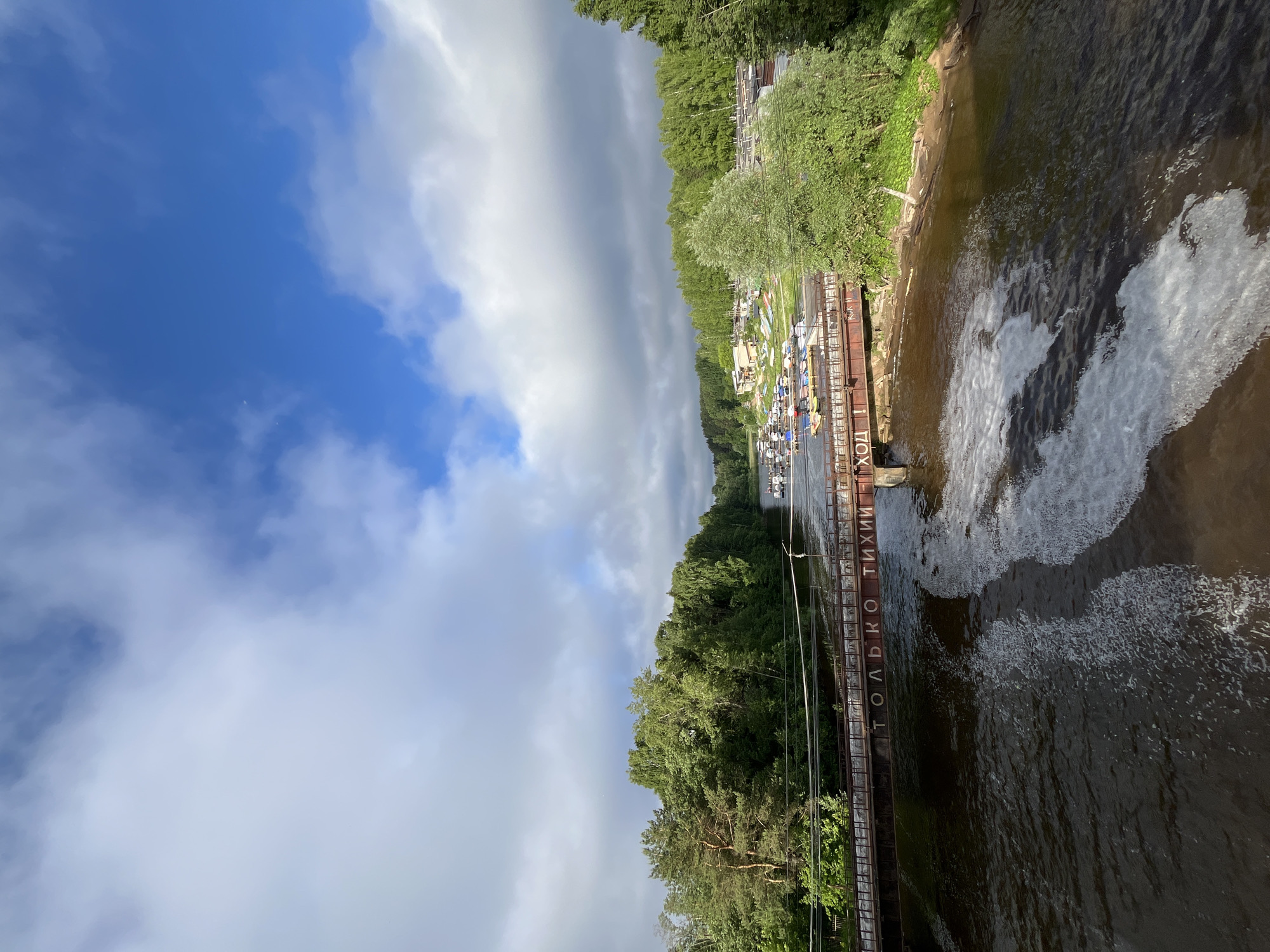 This image captures a scenic view of the Kama River in Chaikovsky, Russia, under a dynamic sky with scattered clouds. A rust-colored metal bridge spans the river, bearing the inscription "ТОЛЬКО ТИХИИ ХОД" (Only quiet passage) in white letters. The water below the bridge exhibits turbulent white foam, suggesting a current or a shallow area.
On the far bank of the river, a lush green forest dominates the landscape, extending along the water's edge. Beyond the trees, a collection of buildings and a marina are visible, with numerous small boats docked. The scene is illuminated by natural daylight, with patches of sunlight breaking through the clouds and casting shadows. No people are discernible in the image. The perspective is from an elevated position, likely another bridge or a vantage point overlooking the river.