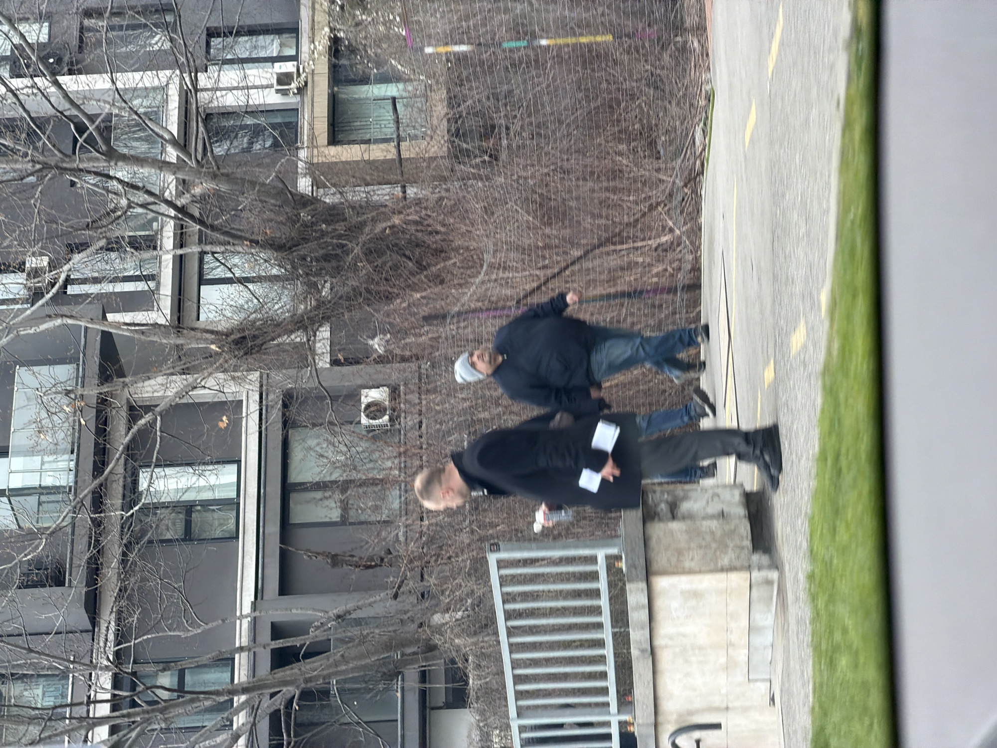 The image depicts two individuals walking on a sidewalk adjacent to a street in what appears to be an urban environment. The sidewalk is marked with yellow lines. To the left of the sidewalk is a building, partially obscured by bare trees. The building has multiple windows and at least one air conditioning unit visible on its facade. One individual is wearing a black jacket and dark trousers, and the other is wearing a dark jacket and blue jeans. The context suggests the location may be in Sofia, Bulgaria, given the architectural style of the building. The image appears to be taken from an elevated position, possibly from a window or balcony.