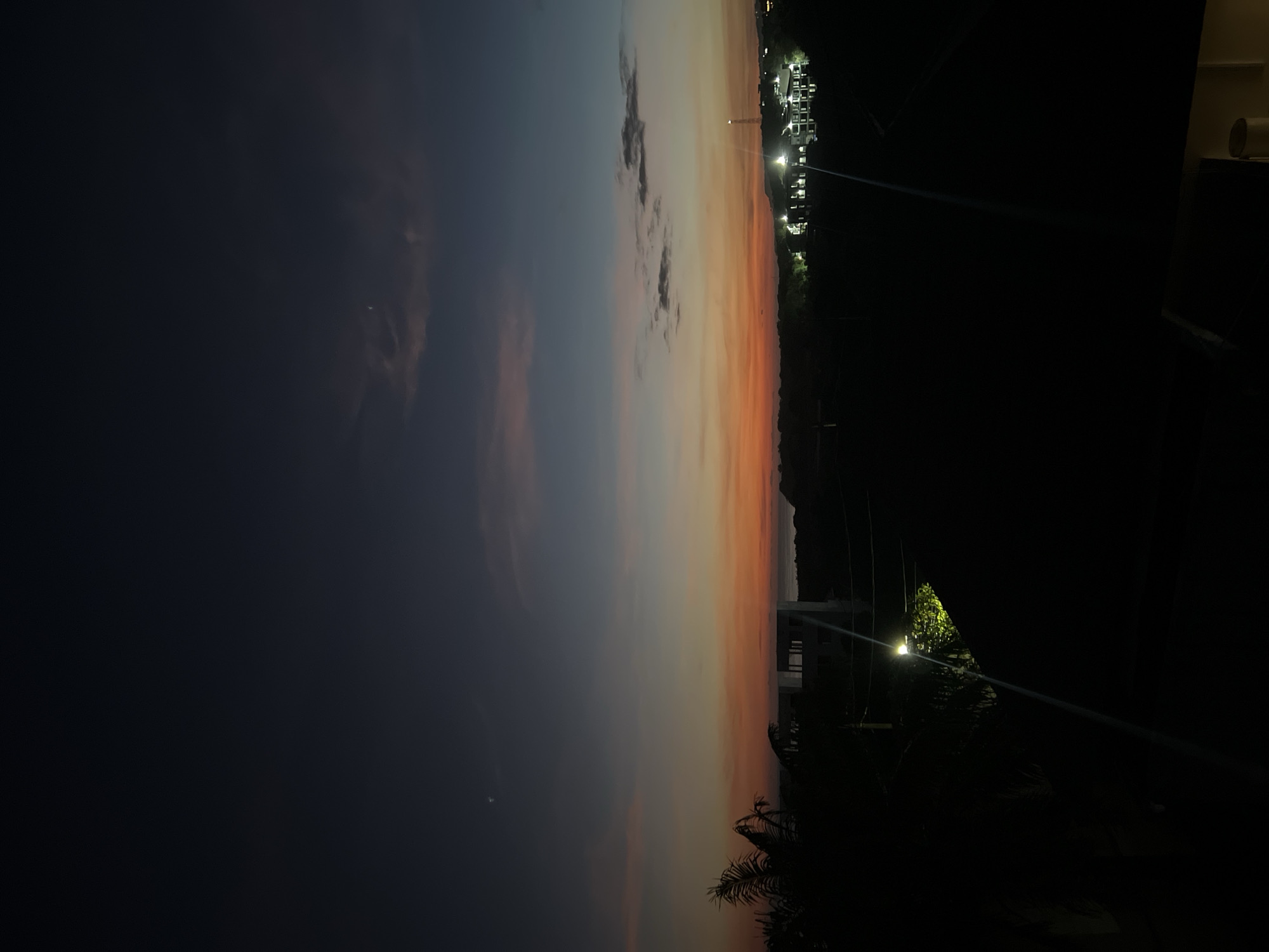 The image captures an outdoor scene at dusk or sunset, oriented vertically in its original format, but describing it as if rotated to its natural landscape orientation. The sky dominates the upper two-thirds of the frame, displaying a gradient of colors transitioning from a deep blue-grey on the left to vibrant oranges, pinks, and yellows on the right, characteristic of twilight. Scattered, soft clouds are visible against this colorful backdrop. A small, bright crescent shape, possibly the moon, is discernible in the darker, upper-left portion of the sky.
Along the horizon line, in the mid-ground, a silhouette of distant buildings or structures is visible, dotted with numerous bright artificial lights, suggesting a populated area or complex. Beyond these structures, faint outlines of hills or mountains stretch across the background.
The foreground is very dark, almost entirely black, indicating deep shadow or lack of illumination. Silhouettes of foliage, possibly palm fronds, are present on the lower right side. A distinct, bright artificial light source, likely a streetlamp, is visible in the lower right, illuminating some unseen greenery nearby. A subtle, horizontal beam of light or lens flare streaks across the mid-ground.
No human figures or specific activities are depicted. The overall impression is one of serene beauty at the end of the day, with the onset of artificial light against the fading natural light. There is no visible text.