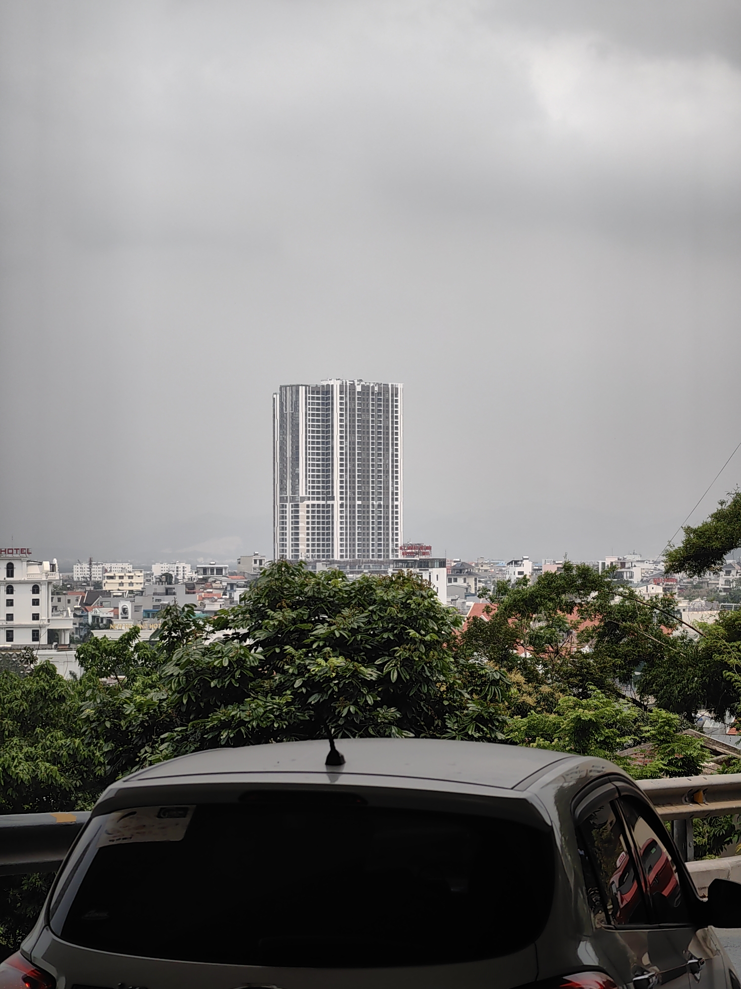 The image depicts a cityscape in Hong Gai Ward, Vietnam, taken from an elevated position. The foreground features the rear of a gray hatchback car, partially obscured by a metal guardrail. Lush green trees fill the middle ground, partially concealing rooftops of buildings. In the distance, a tall, modern skyscraper with white facade stands prominently against an overcast, gray sky. Smaller buildings, including a hotel with visible signage, are visible below the skyscraper. The overall atmosphere is muted due to the weather conditions.