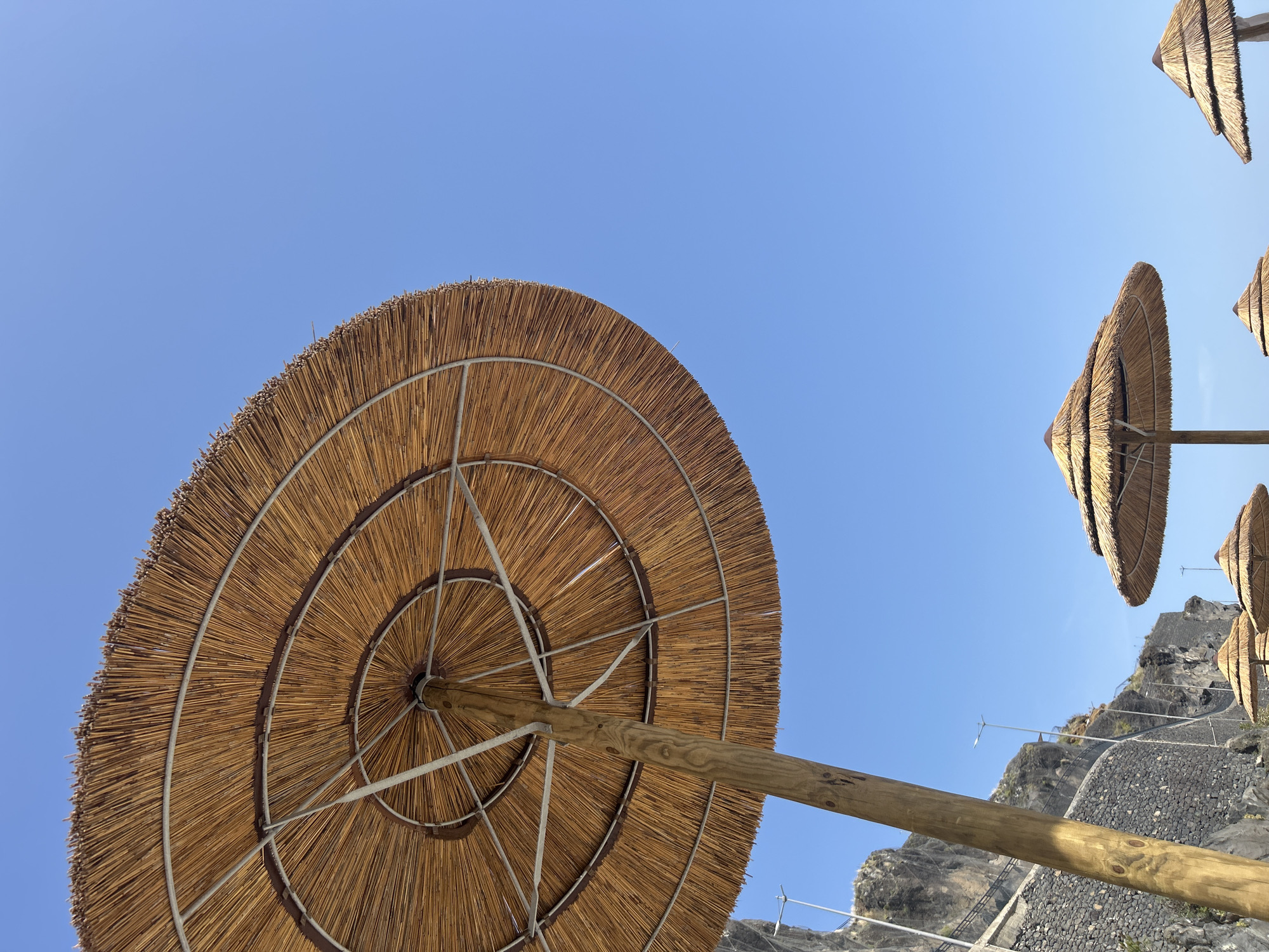 The image features a low-angle view of a beach with straw parasols against a clear blue sky. The focus is on the underside of one parasol, revealing its wooden support structure and the concentric rings of straw that form its canopy. Several other parasols are visible in the background. The terrain appears rocky and mountainous, with stone walls and staircases evident. The picture shows this area, likely in Câmara de Lobos, Portugal.