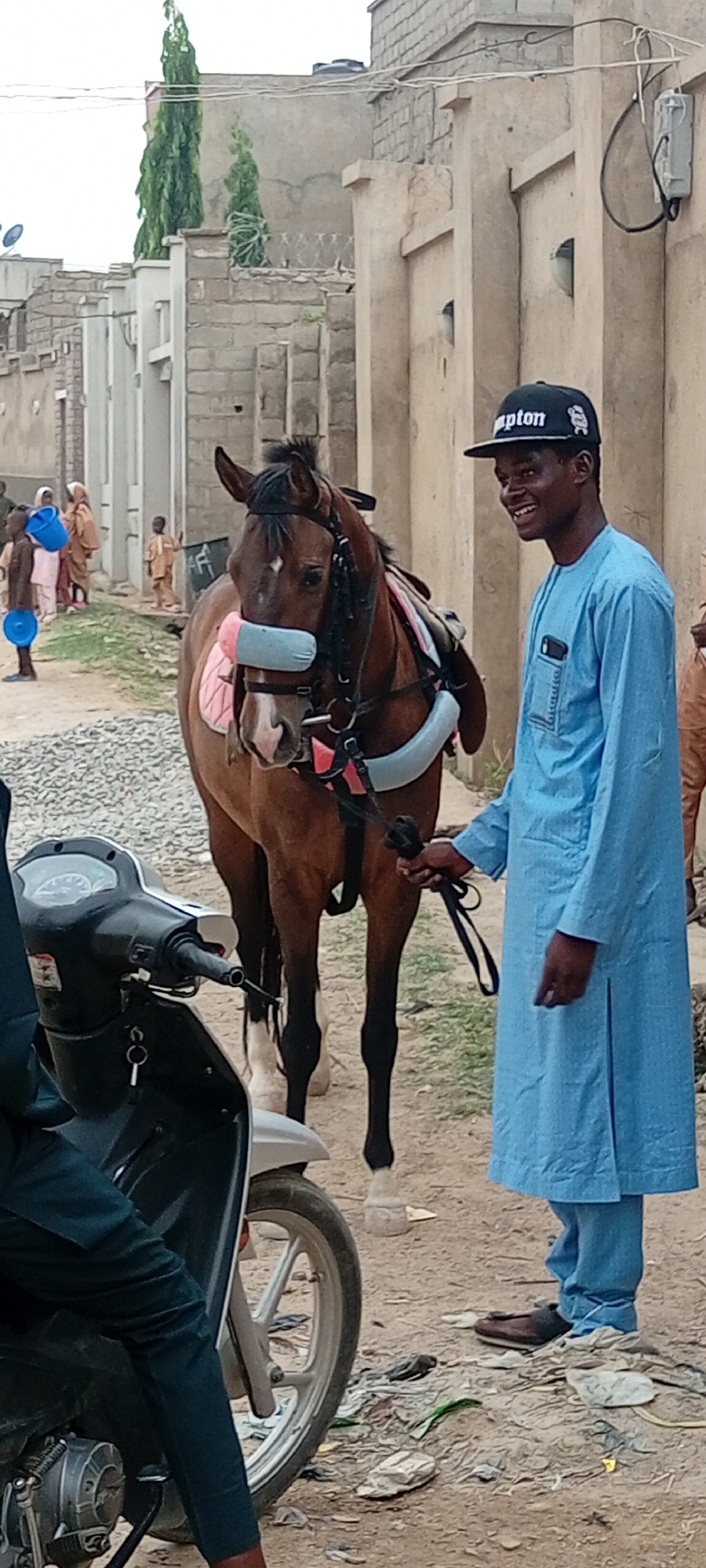 The image shows a man and a horse standing on a dirt street in a city identified as Unknown. The man, who is smiling and wearing a light blue robe and a "Compton" baseball cap, holds the horse's reins. The horse is saddled with blue padding. In the foreground, a partially visible scooter is parked. In the background, blurred figures suggest a small group of people, and buildings line the street. The scene appears to be an everyday occurrence in an urban or semi-urban location.